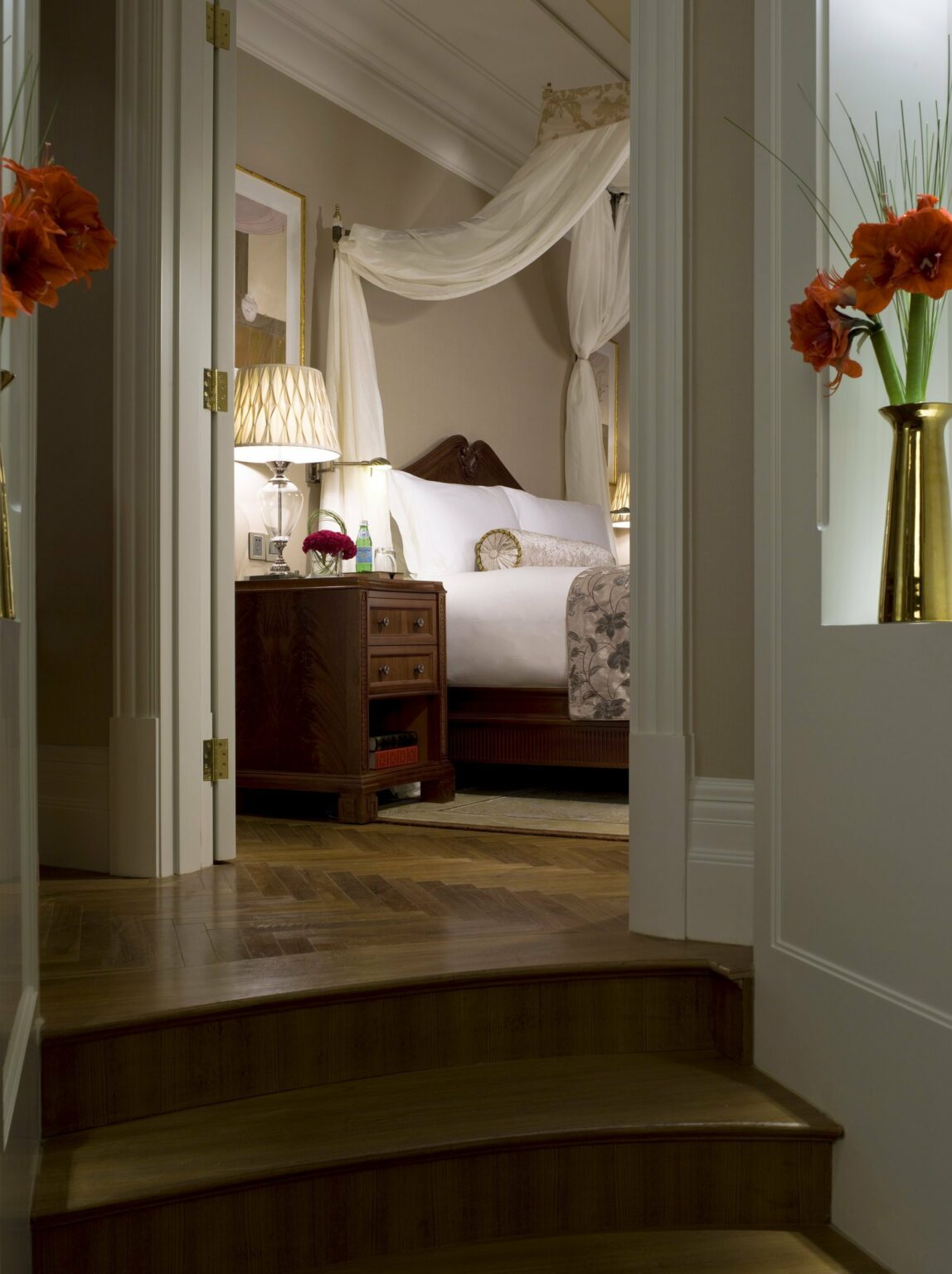 Cozy bedroom view through a staircase doorway, featuring a canopy bed with white linens, a wooden nightstand with a lamp, and an elegant, calm ambiance.