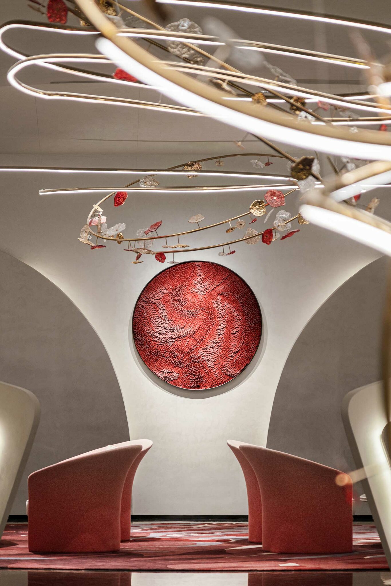 Modern interior with two curved, pink chairs facing a circular, textured red artwork on a white wall. Overhead is an intricate, artistic light fixture.