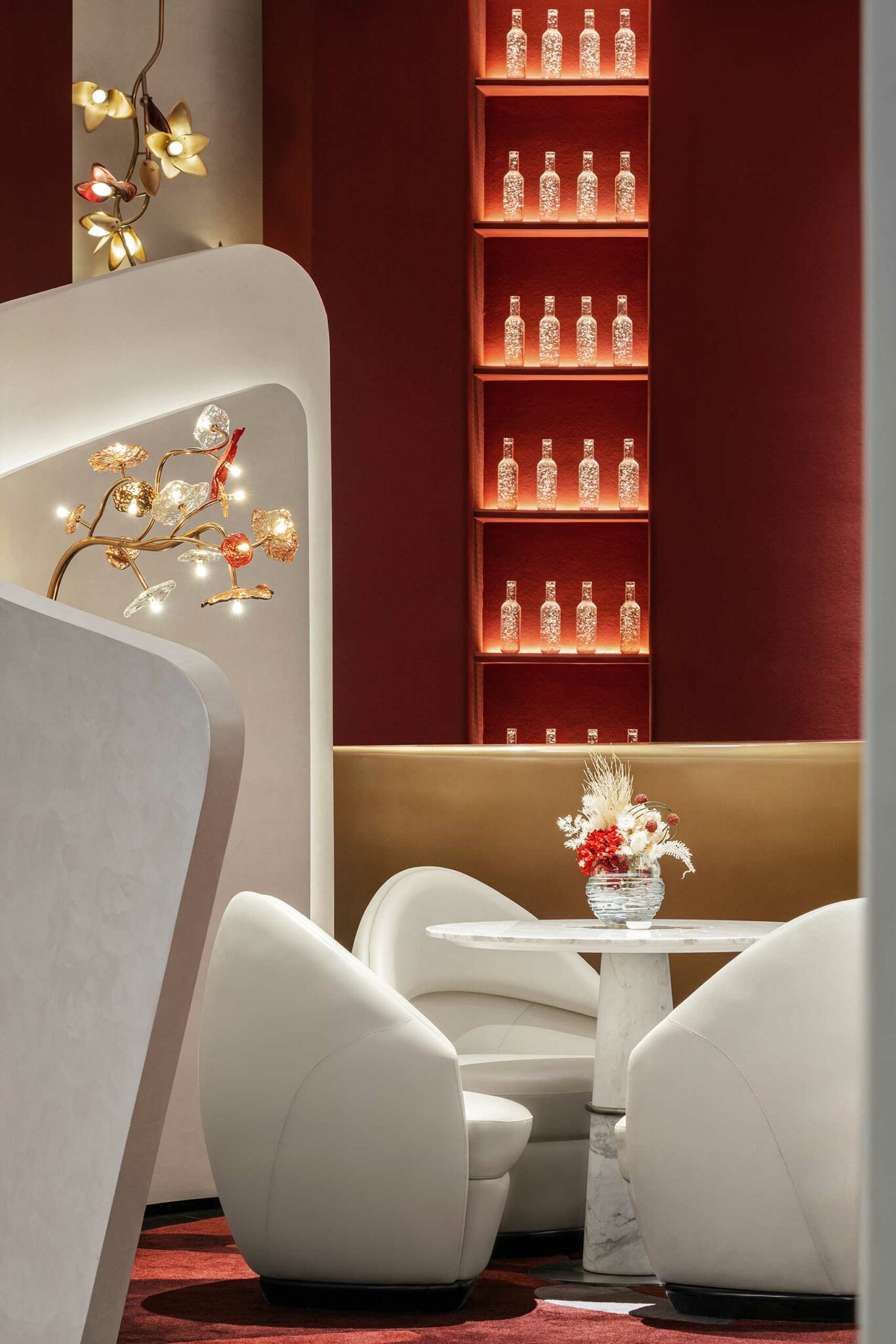 A modern, elegant scene of a workplace lounge with a marble table, white chairs, and a floral centerpiece. The red wall features shelves with decorative glass bottles. Warm lighting and floral lamp accents add a cozy ambiance.