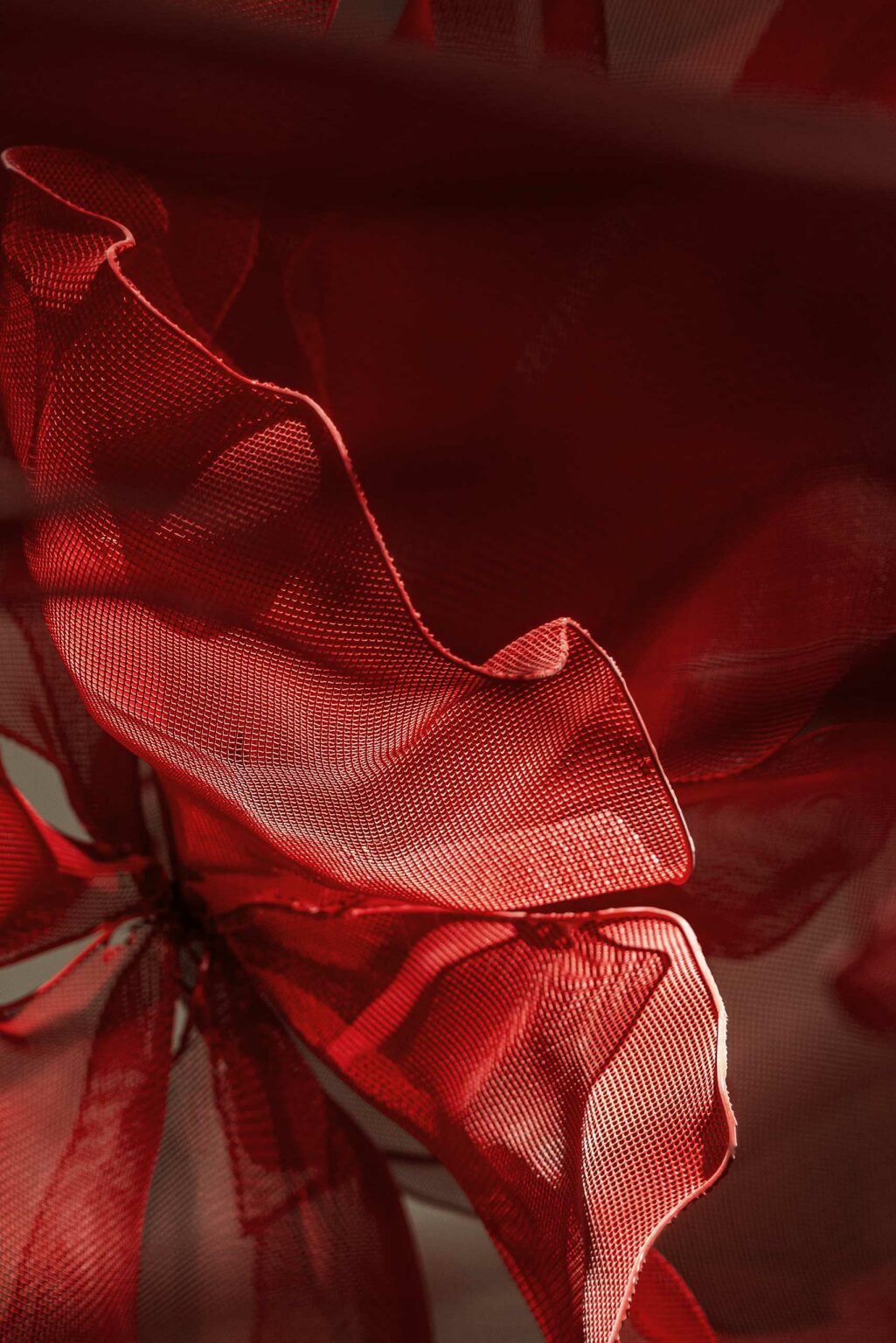 Close-up of a flowing red mesh fabric with soft, curved folds. The texture is light and airy, creating a dynamic and artistic appearance.