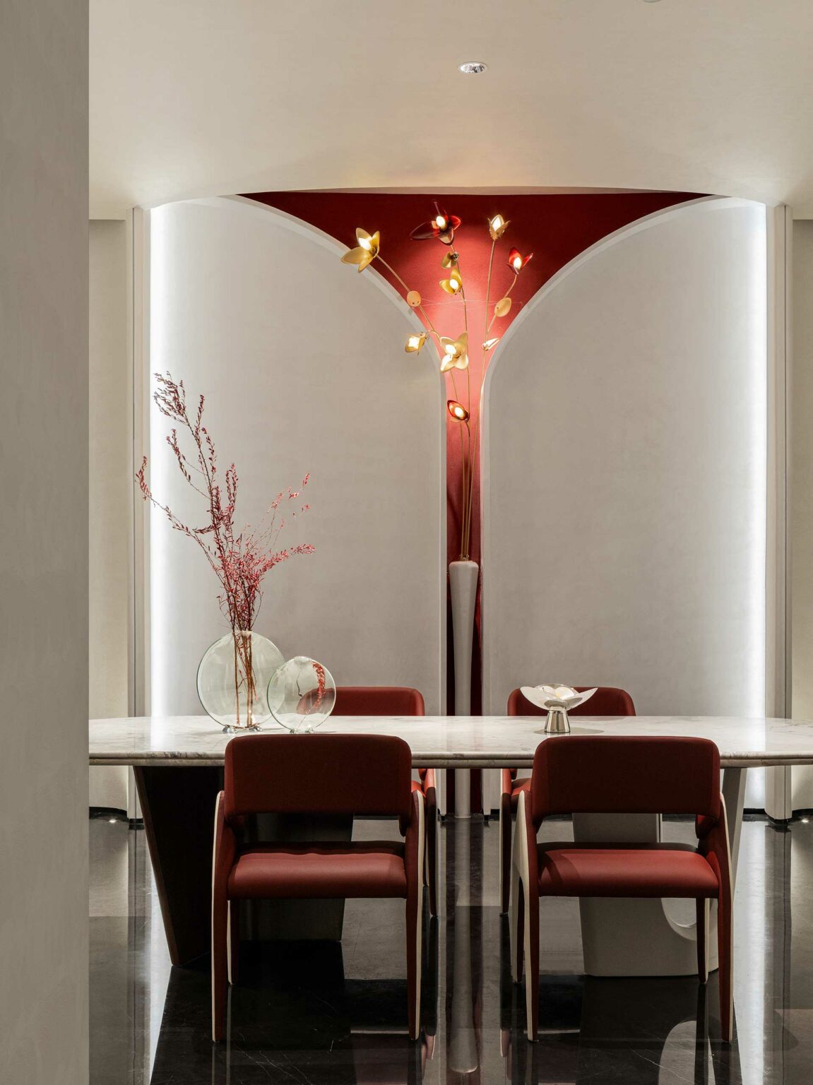 Modern dining area with a marble table, four red chairs, and decorative wall lights resembling flowers. A vase with red branches adds elegance.