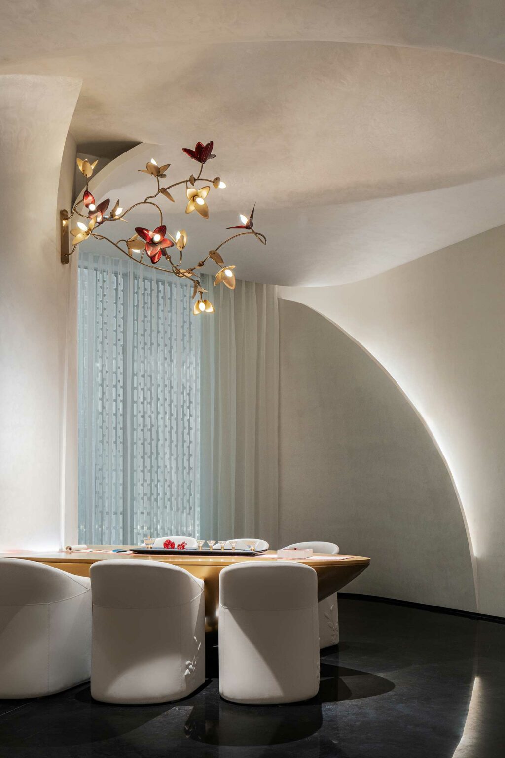 Elegant dining room with white walls and plush chairs around a sleek table. A sculptural light fixture with red flower accents hangs above, creating a modern, serene atmosphere.