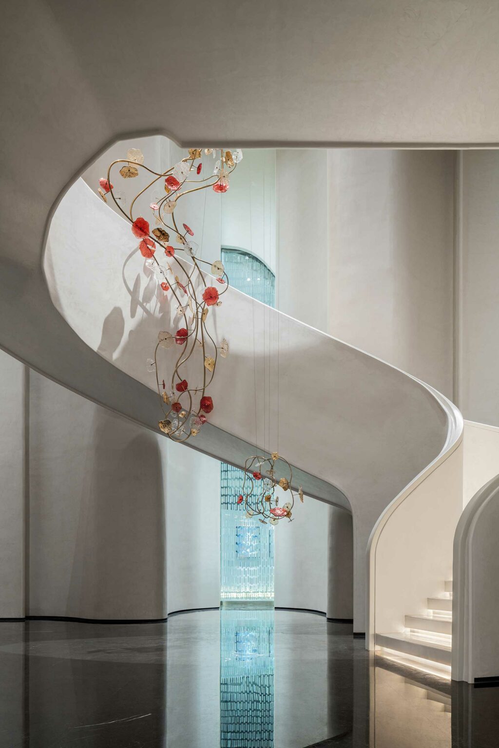 A curved white staircase with a floral chandelier featuring red and gold elements hangs elegantly. The setting feels modern and spacious with reflective flooring.