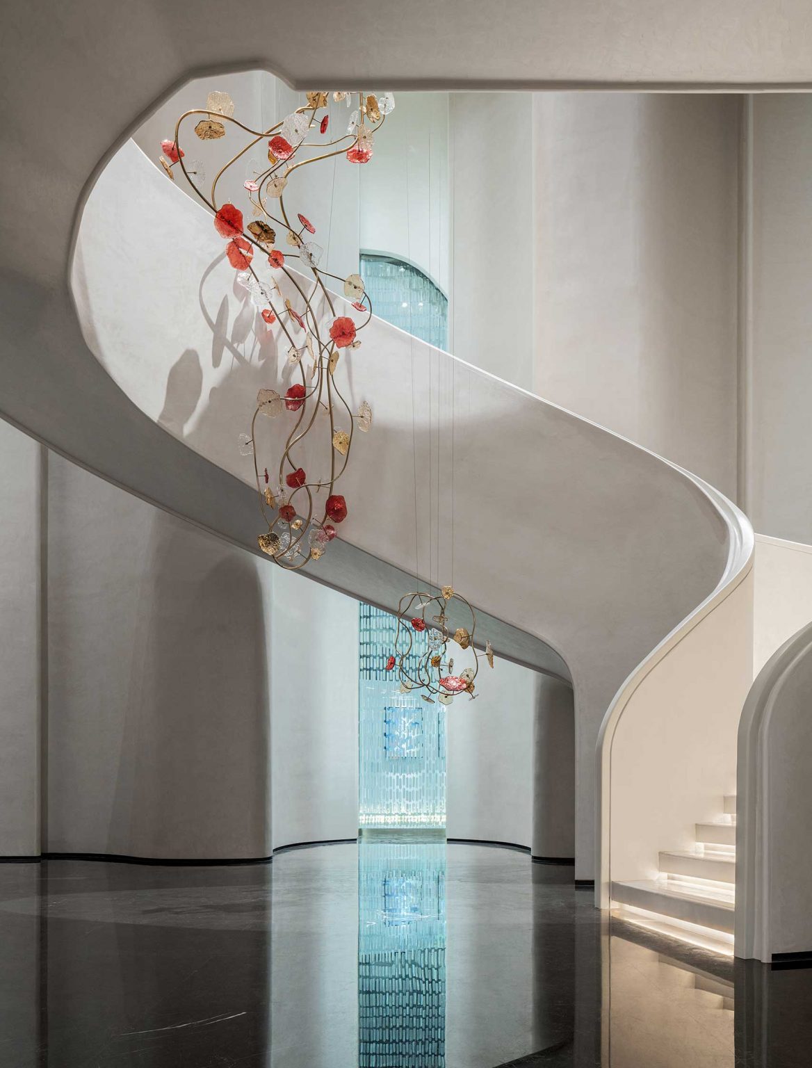 A curved white staircase with a floral chandelier featuring red and gold elements hangs elegantly. The setting feels modern and spacious with reflective flooring.