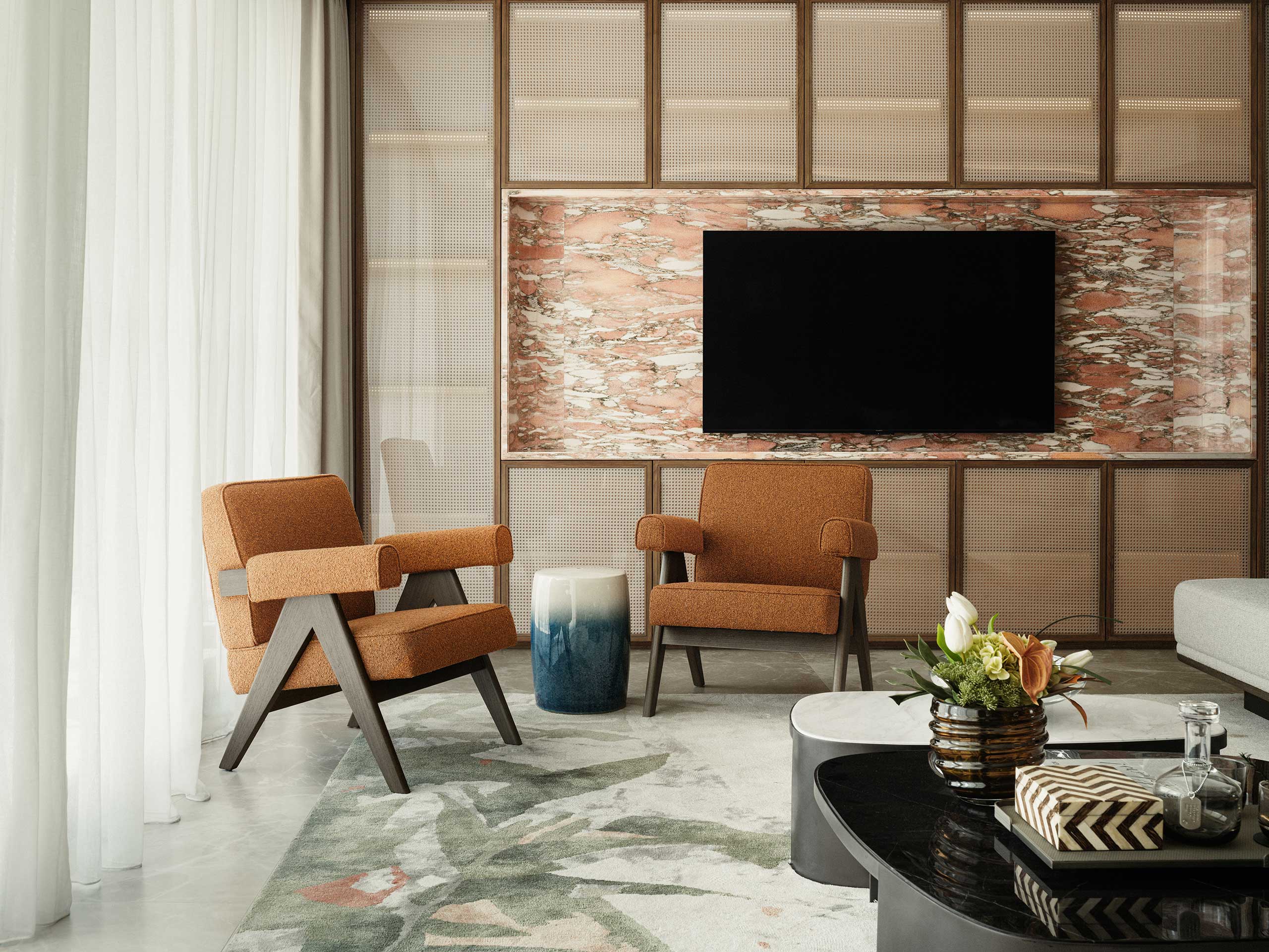 Contemporary living room with two orange armchairs, a blue and white stool, abstract rug, and large wall-mounted TV. Neutral tones evoke a calm atmosphere.
