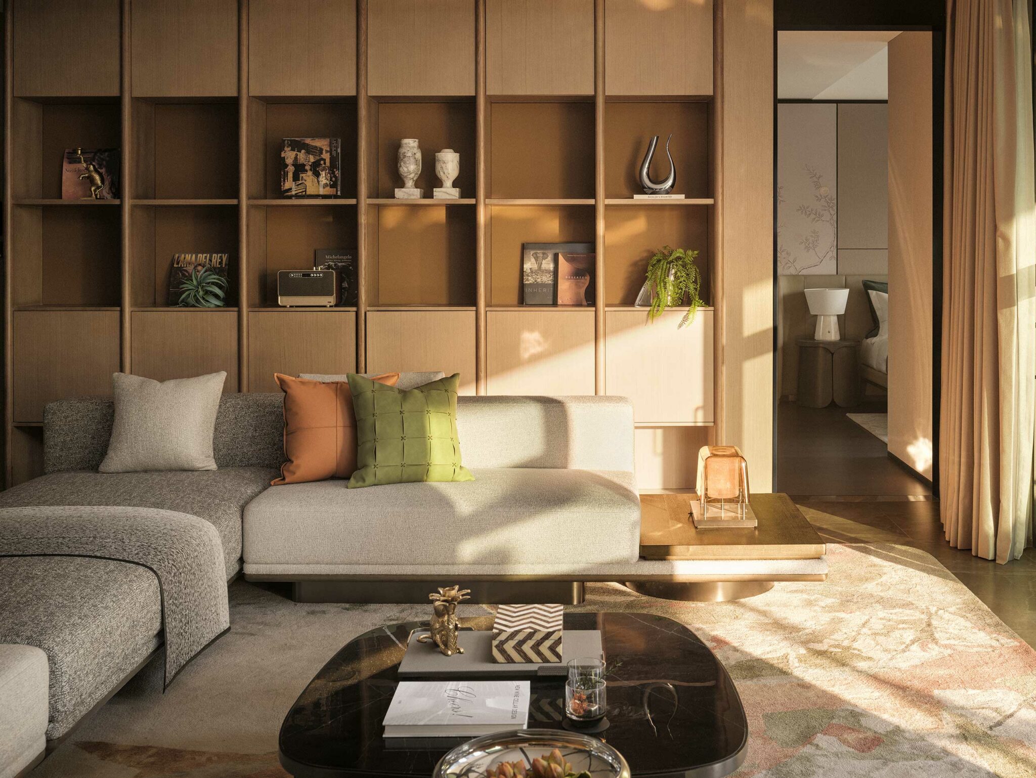Modern living room with a gray sectional sofa accented with green and orange pillows. Sunlit wooden shelves display decor. Calm, cozy ambiance.