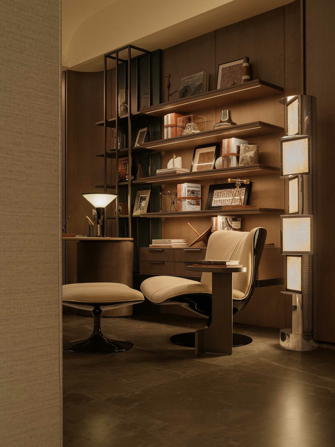 A cozy, elegant study with dim lighting features a sleek modern chair and ottoman, wooden shelves with books, and decorative items. Warm tones create a peaceful ambiance.