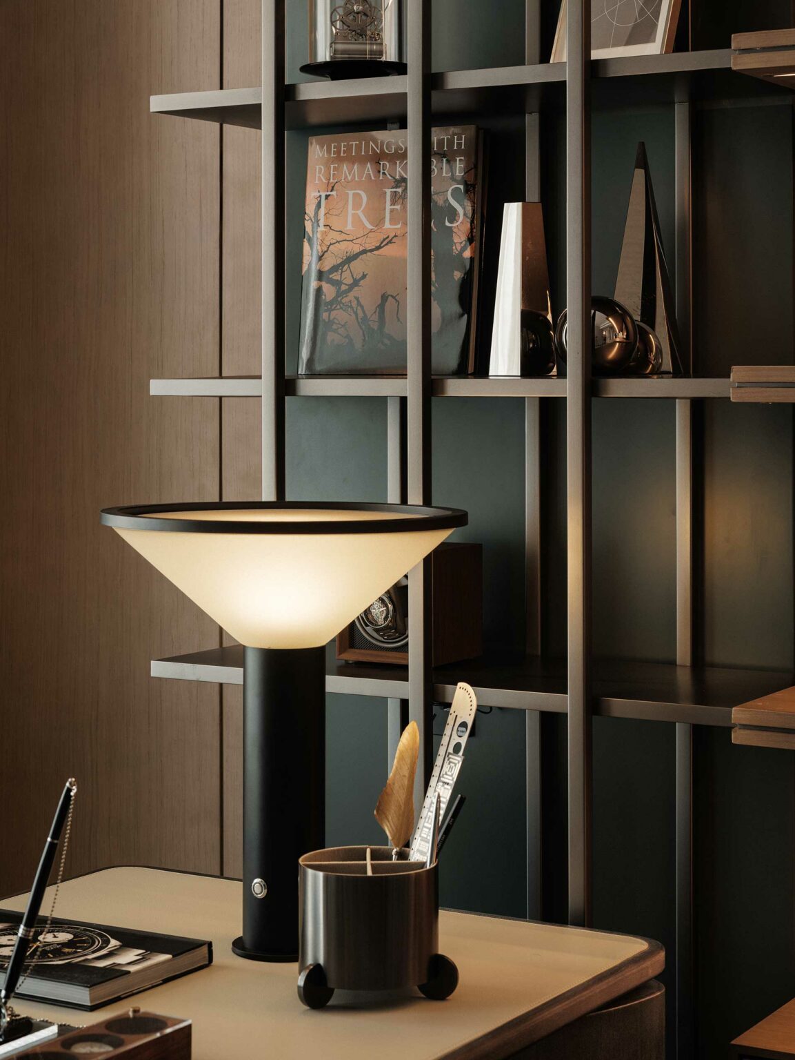 A stylish desk setup featuring a modern lamp, a sleek container for utensils, and a bookshelf with decorative items and books, creating a sophisticated and functional workspace.