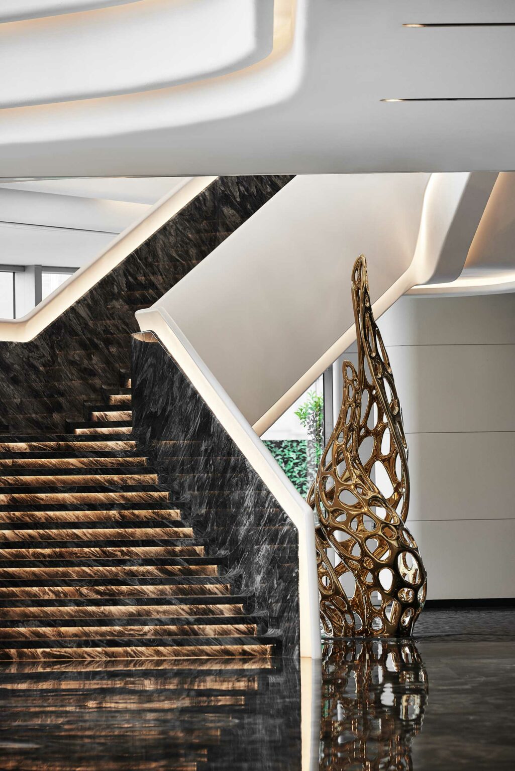 A sleek, modern staircase in a office lobby setting with dark marble steps and white railings leads upward. Beside it, an abstract golden sculpture stands on a glossy floor.