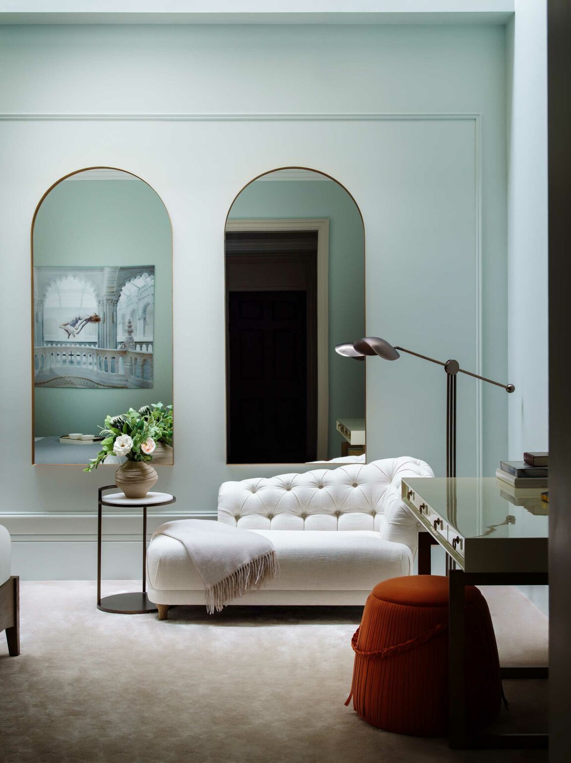 Elegant living room featuring a white chaise lounge with a throw, a side table with a floral arrangement, and twin arch mirrors reflecting light, creating a serene atmosphere.