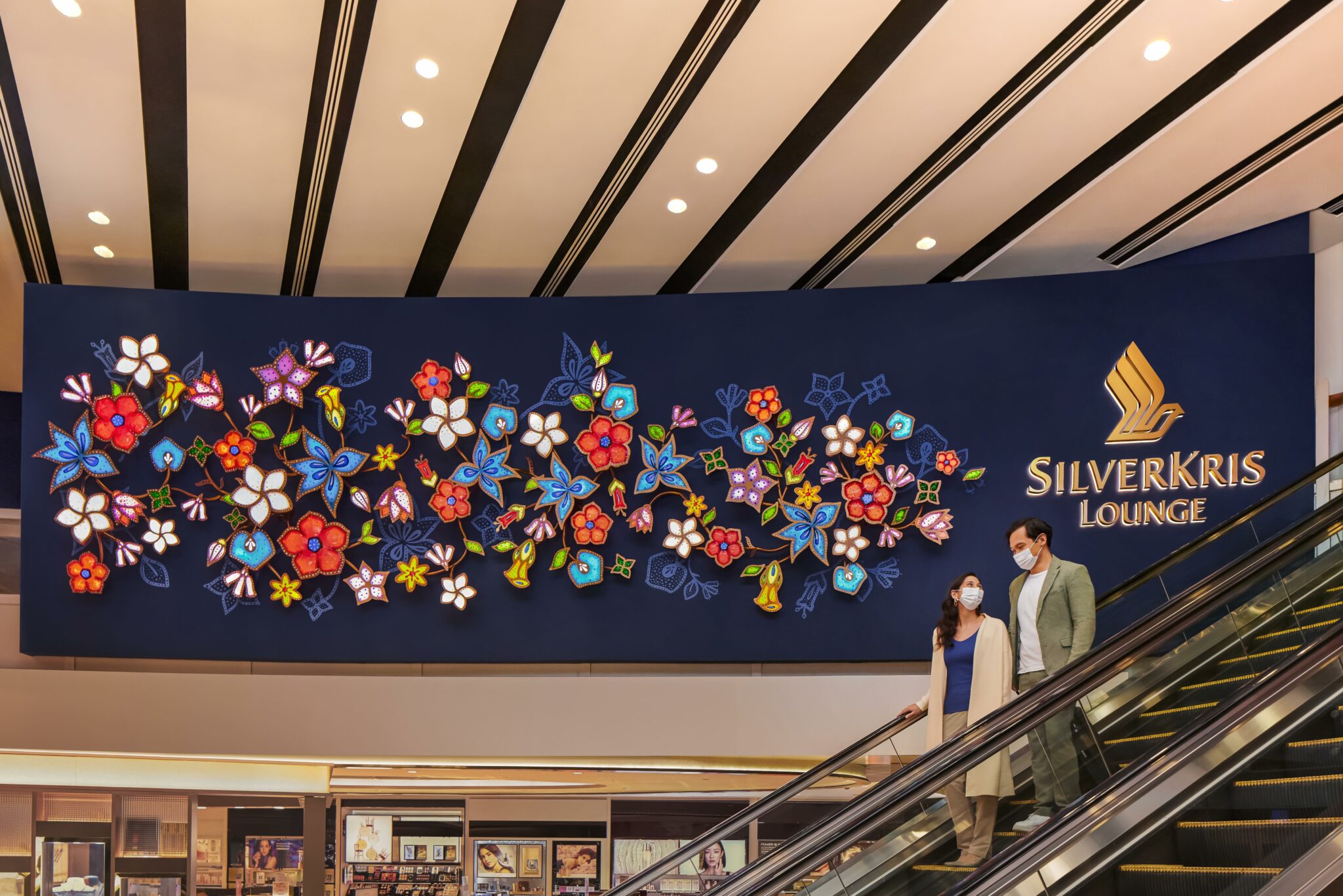 Guests descent an escalator toward Singapore Airlines SilverKris Lounge, featuring a vibrant floral mural and elegant branding, creating a welcoming and stylish entryway into a luxurious lounge environment.