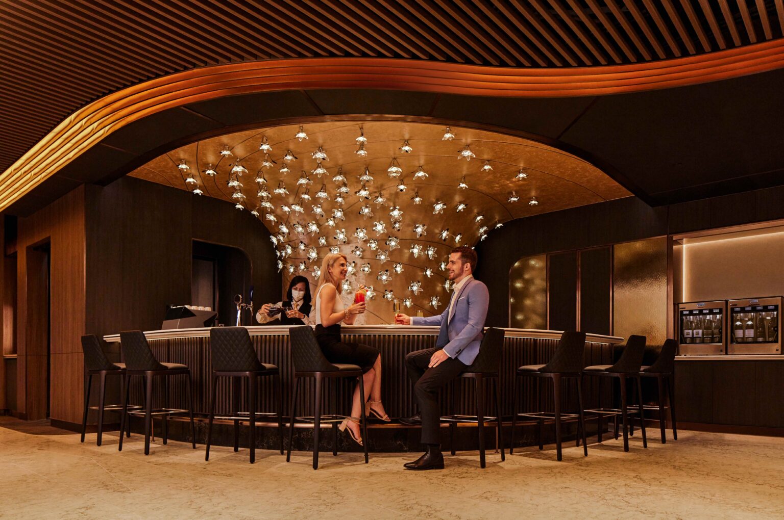 A chic airline bar scene featuring a couple enjoying drinks at a stylish counter, illuminated by a unique chandelier, creating a vibrant and upscale atmosphere for socializing and relaxation.