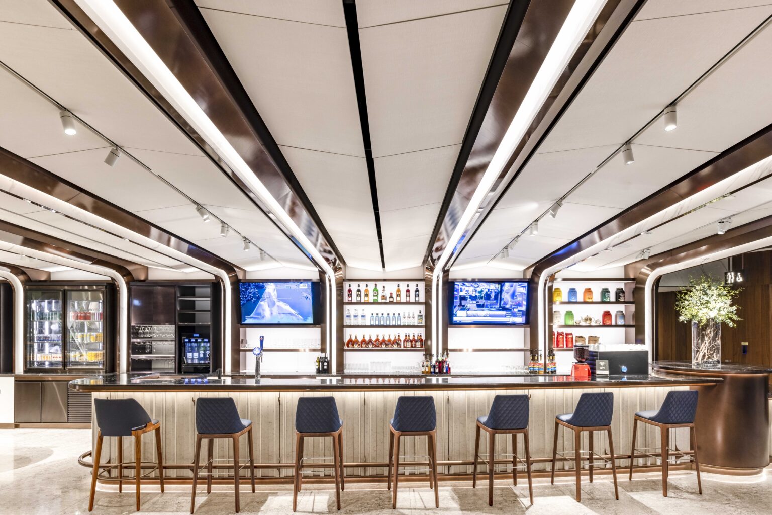 A modern bar with sleek design, featuring high stools, illuminated shelves with beverages, and multiple screens, creating an inviting space for guests to enjoy drinks and entertainment.