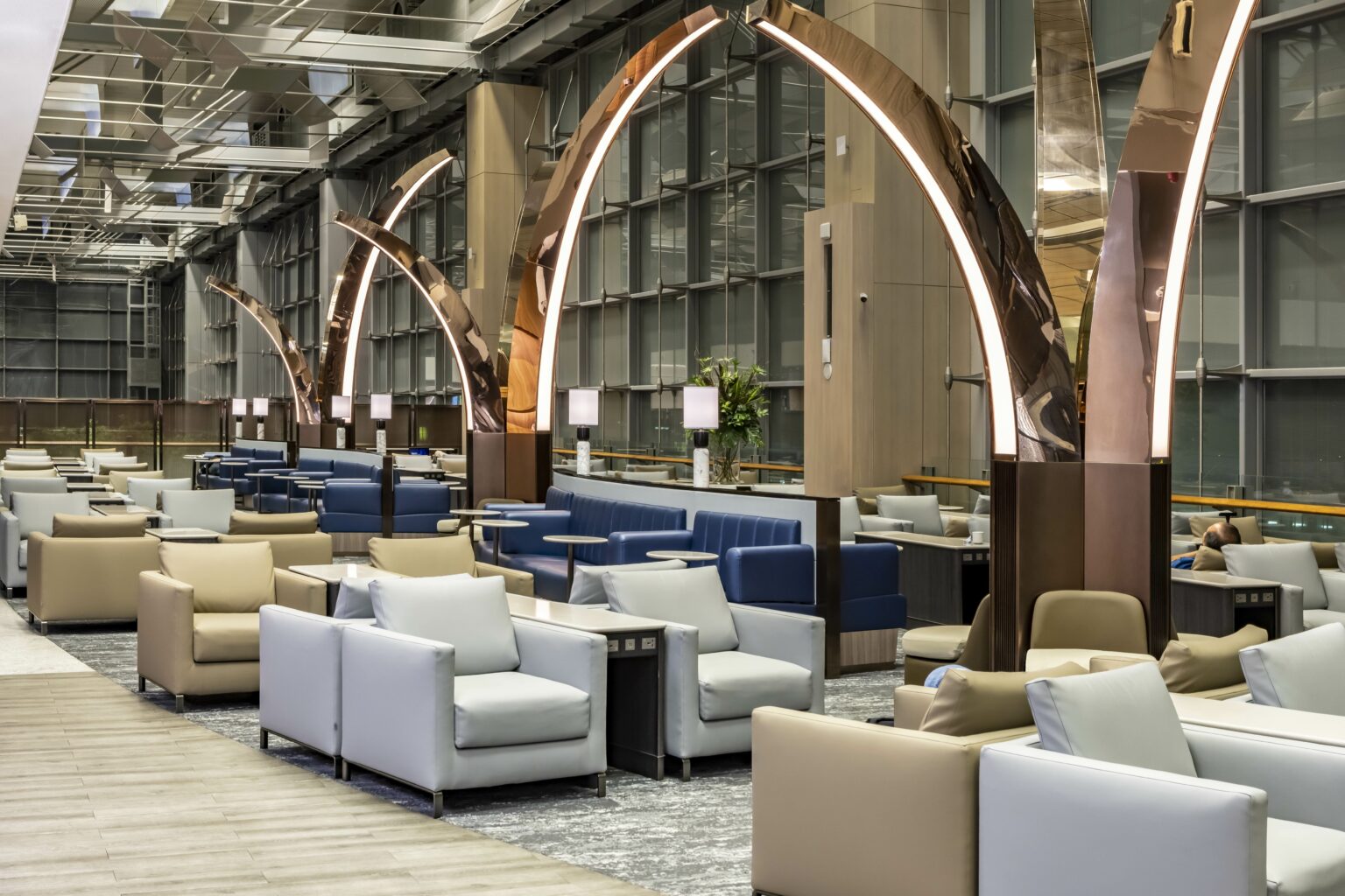 A spacious airline lounge area featuring stylish blue and white seating, elegant arches with ambient lighting, and large windows, creating a bright and inviting atmosphere for relaxation and socializing.