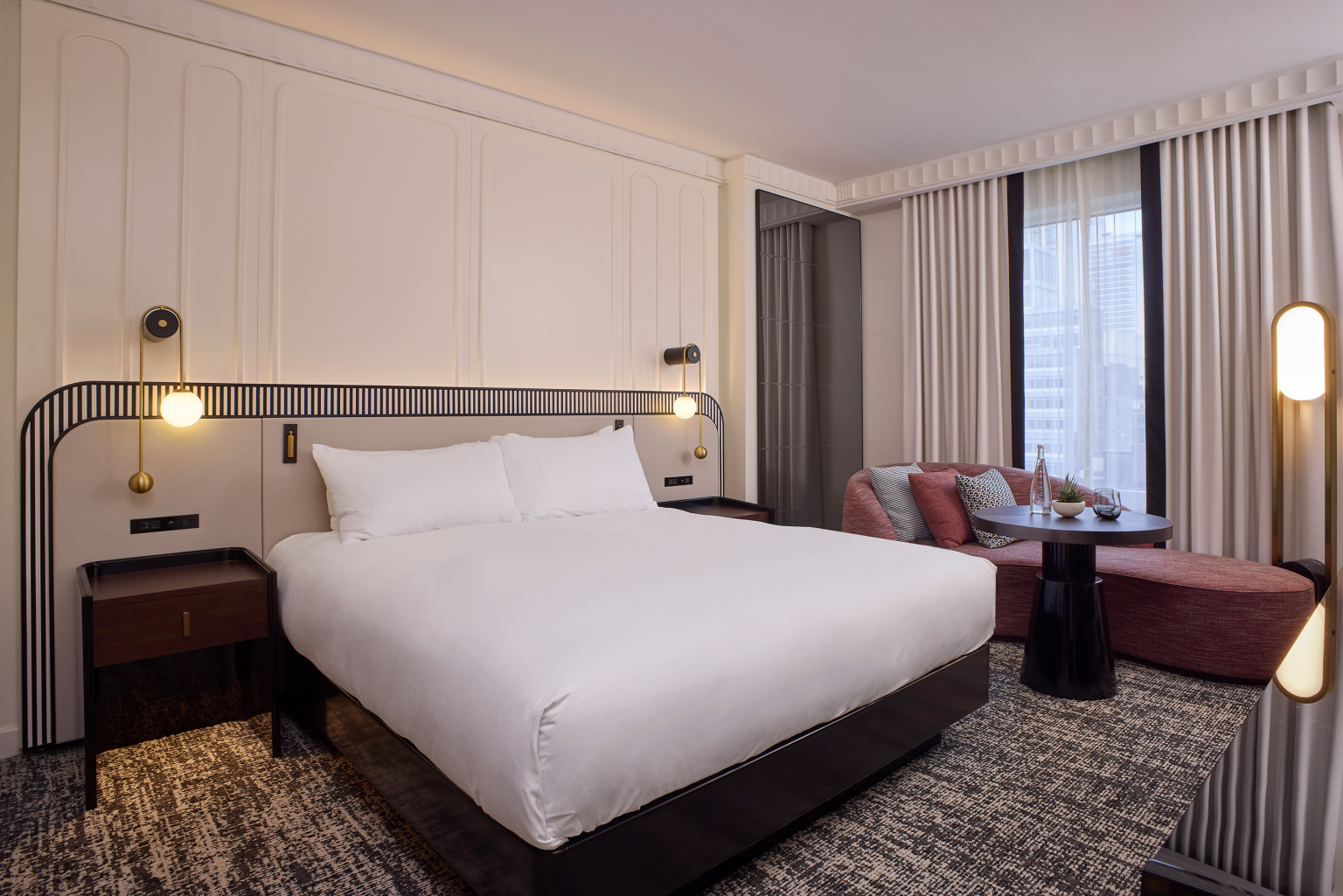 Modern hotel room with a plush white bed, bedside lights, and nightstands. Cozy seating area with red sofa and pillows near large, curtained windows.