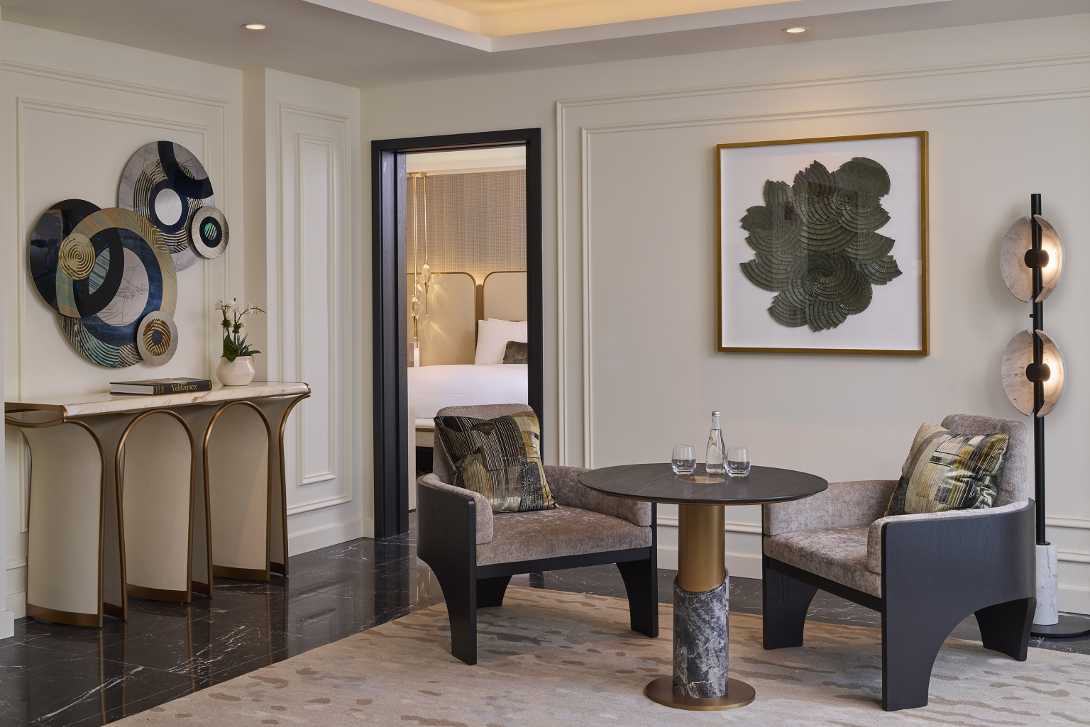 Elegant hotel suite lounge with modern art on walls, two plush chairs around a small round table, and a doorway leading to a bedroom. Cozy and luxurious ambiance.