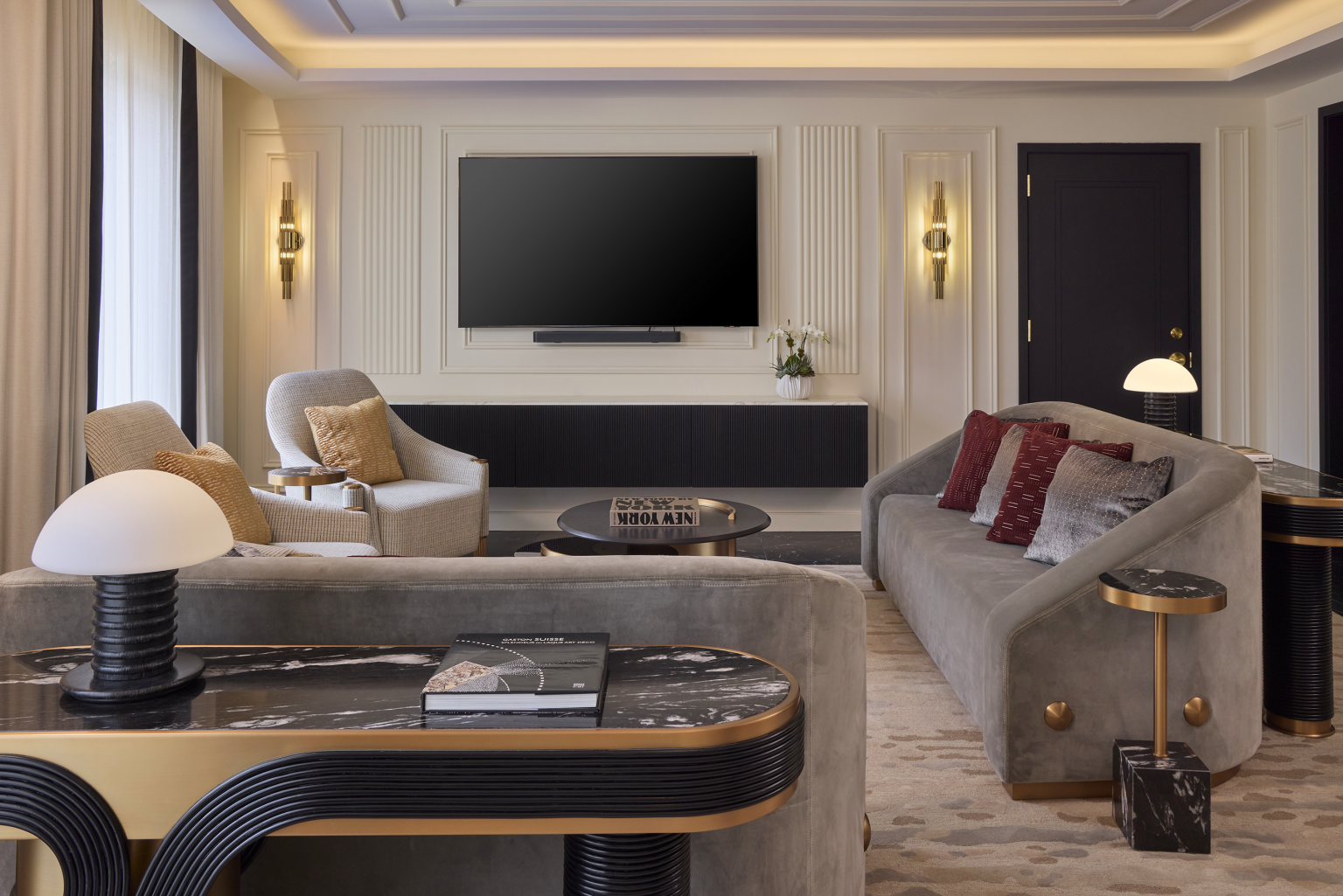 Luxurious living room with a large TV, chic gray sofas adorned with red and gray pillows. Soft lighting, stylish decor, and marble tables create an elegant ambiance.