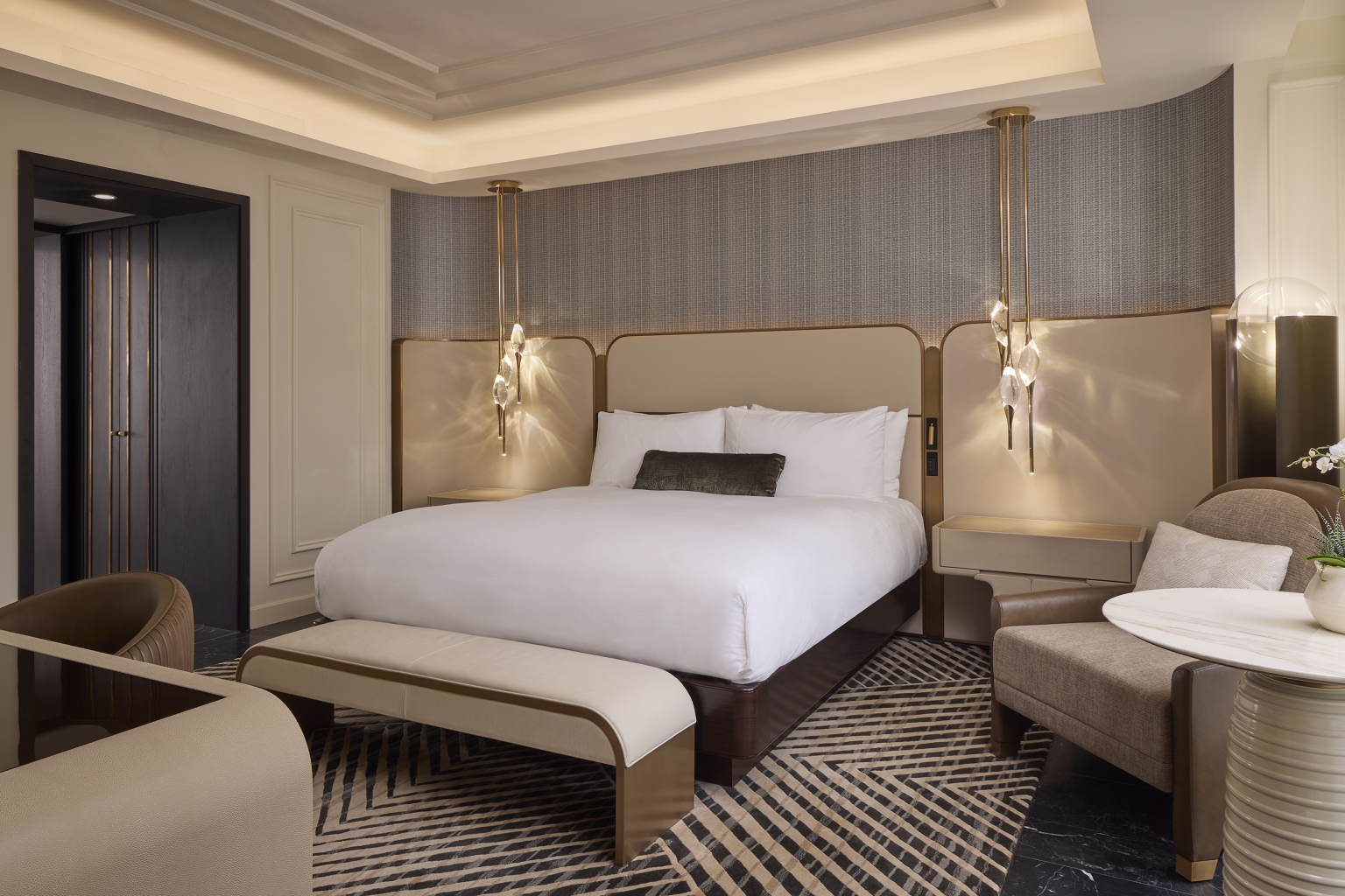 Luxurious hotel room with a large bed, elegant lighting, and a modern design. Features a cozy seating area, geometric rug, and neutral tones.