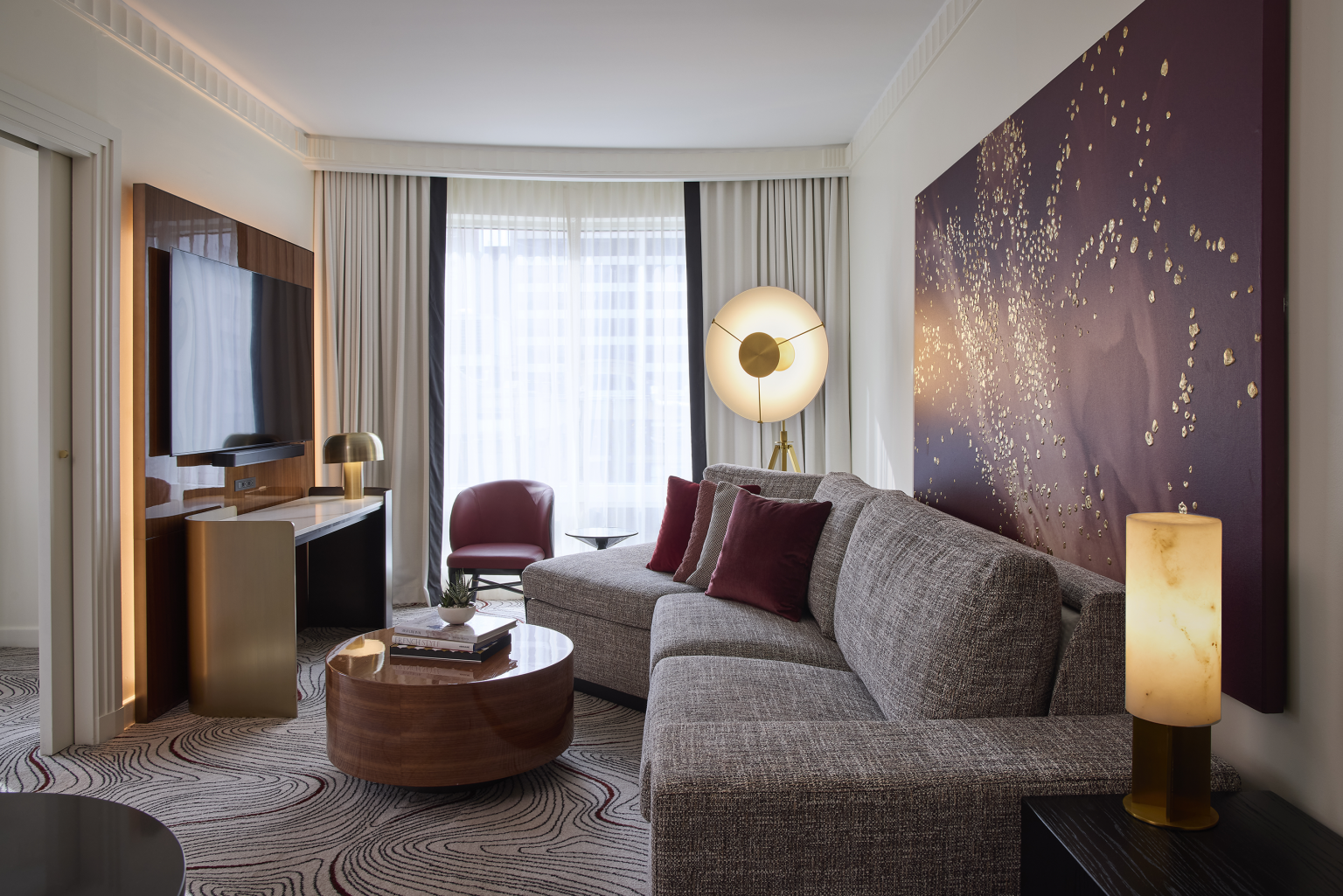 Elegant hotel room with a modern gray sofa, red cushions, chic lighting, and a large abstract art piece. Cozy ambiance with soft lighting and stylish decor.