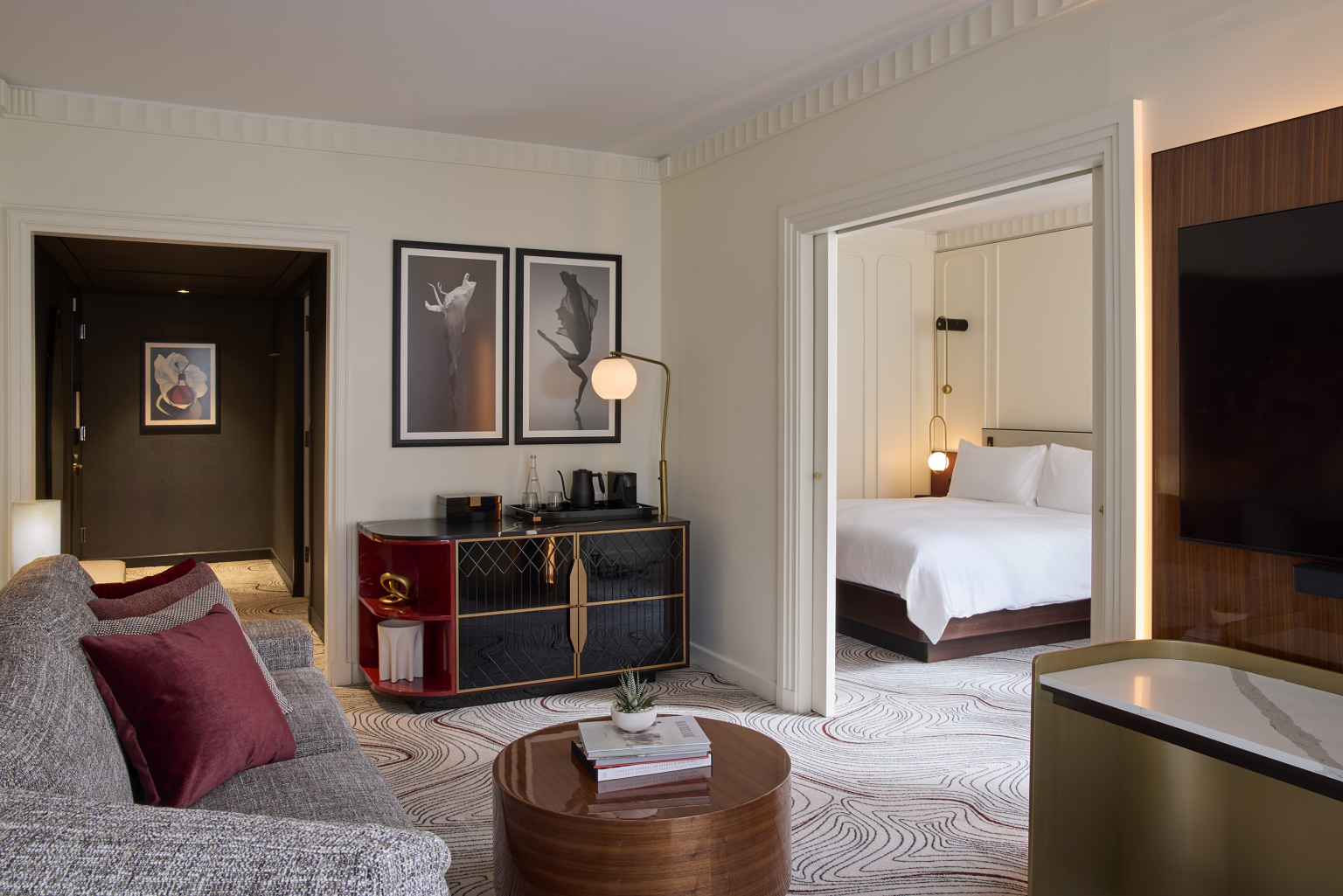 Modern hotel suite with a living area and bedroom. A gray sofa with red pillows, abstract art, and round wooden coffee table create a cozy ambiance.