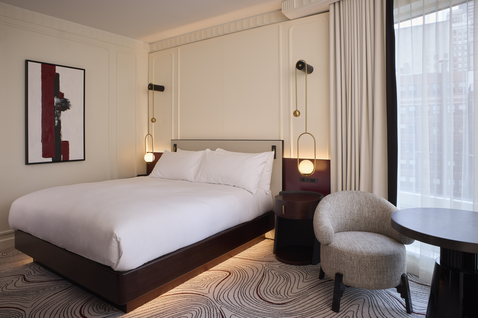 Modern hotel room with a large bed, white bedding, abstract wall art, and elegant lighting. Cozy armchair and table by a curtained window, creating a serene ambiance.
