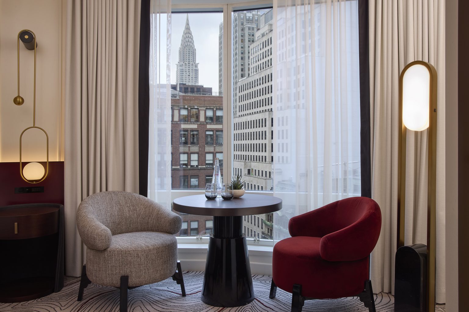 Cozy hotel room with a round table, two modern chairs—grey and red—by a large window showcasing city buildings. Relaxed, urban ambiance.