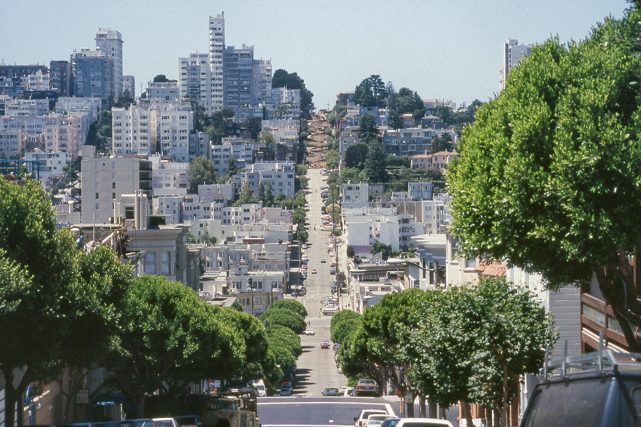 Steep street in San Francisco, lined with lush green trees and surrounded by modern high-rise and mid-rise buildings under a clear blue sky.