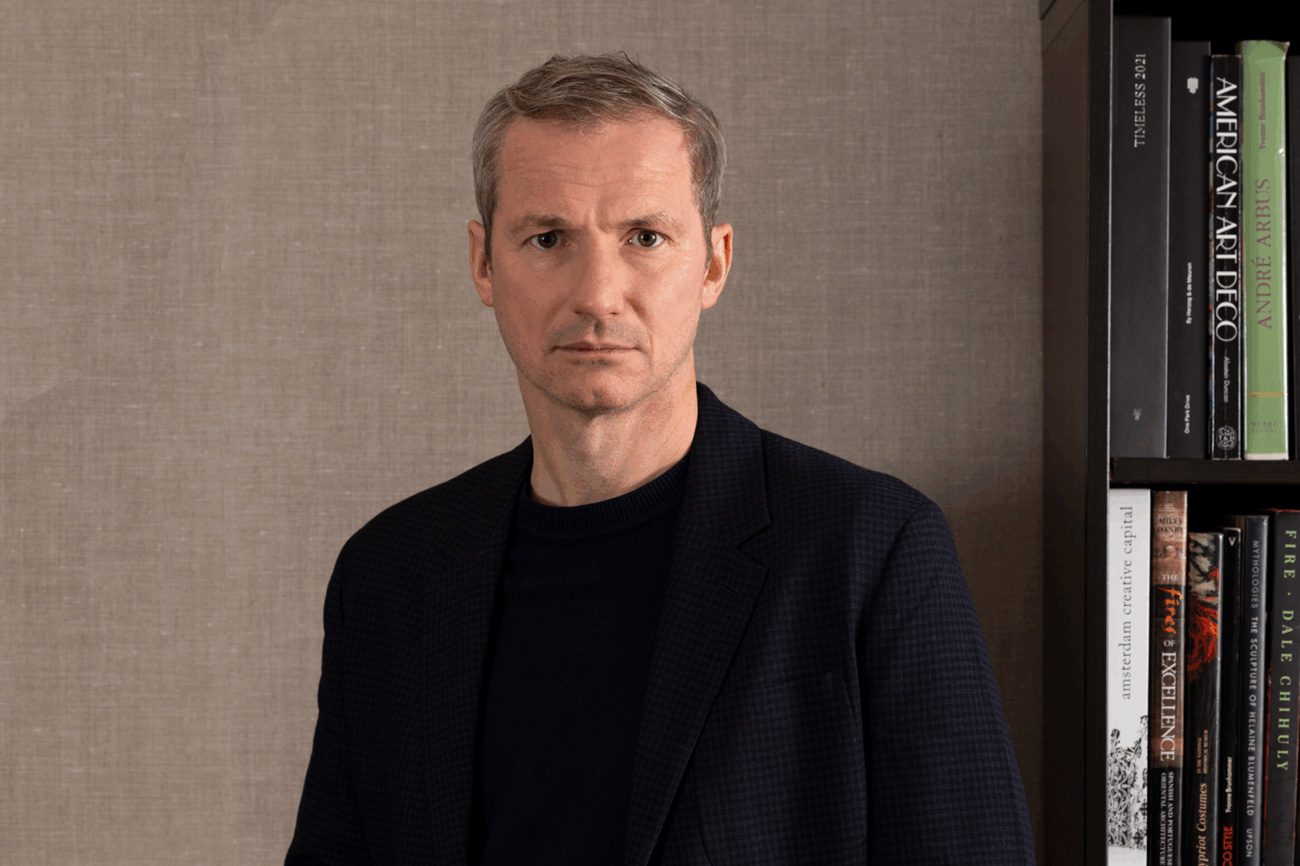Portrait of HBA Co-CEO Chris Godfrey wearing a dark blazer and sweater standing against a textured beige backdrop, looking serious and contemplative.