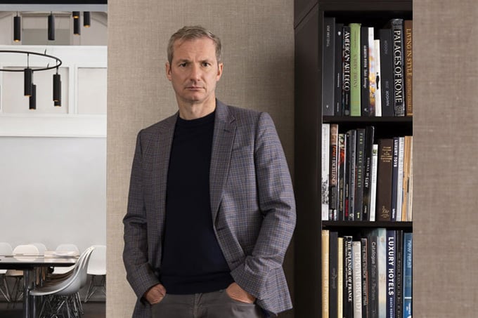Portrait of HBA Co-CEO Chris Godfrey wearing a dark blazer and sweater standing against a home library backdrop, looking serious and contemplative.