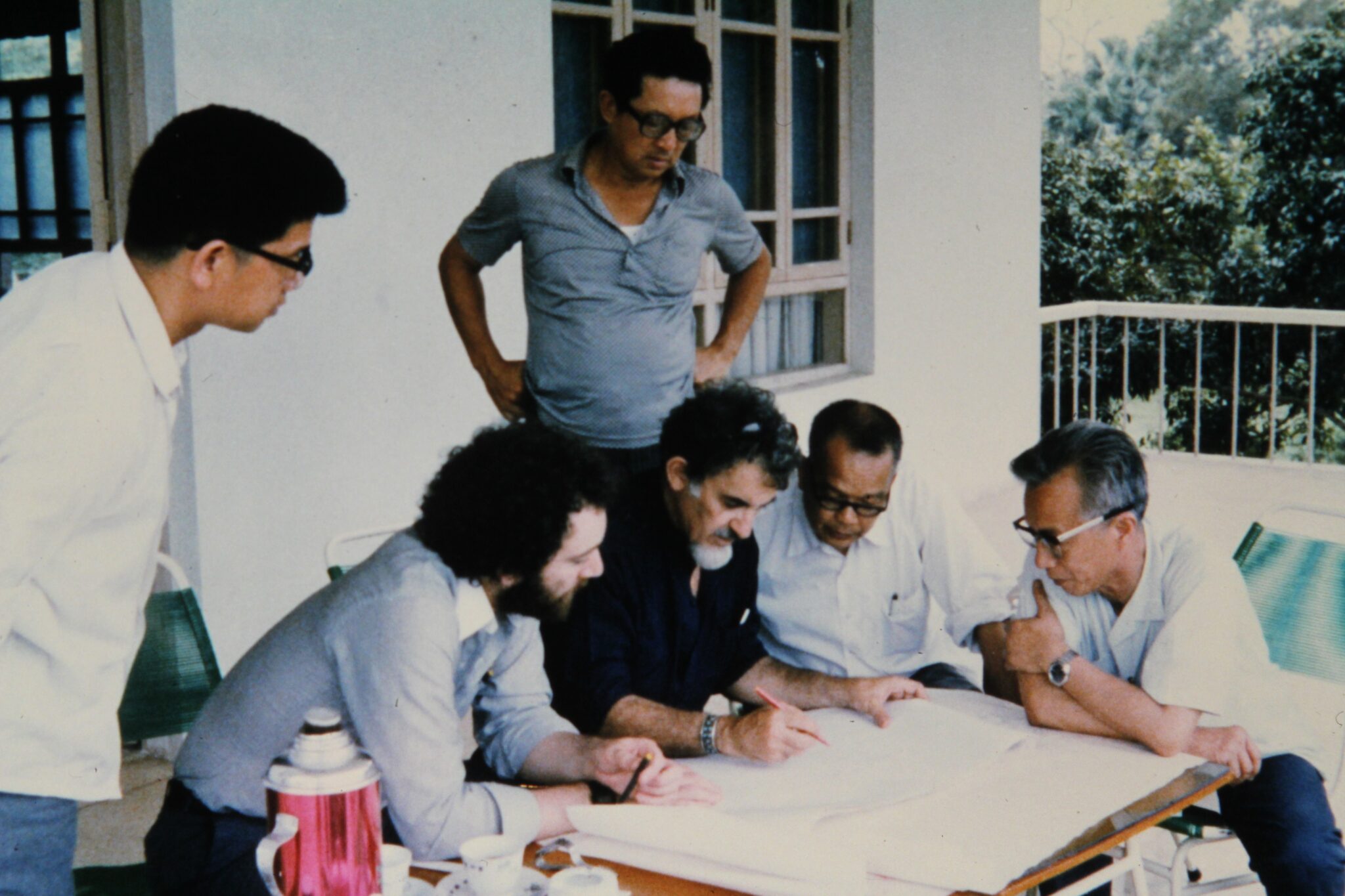 Six men gather around a table on a porch, intently discussing documents about HBA's first-ever project in China. One man writes while others observe. Trees are visible in the background.