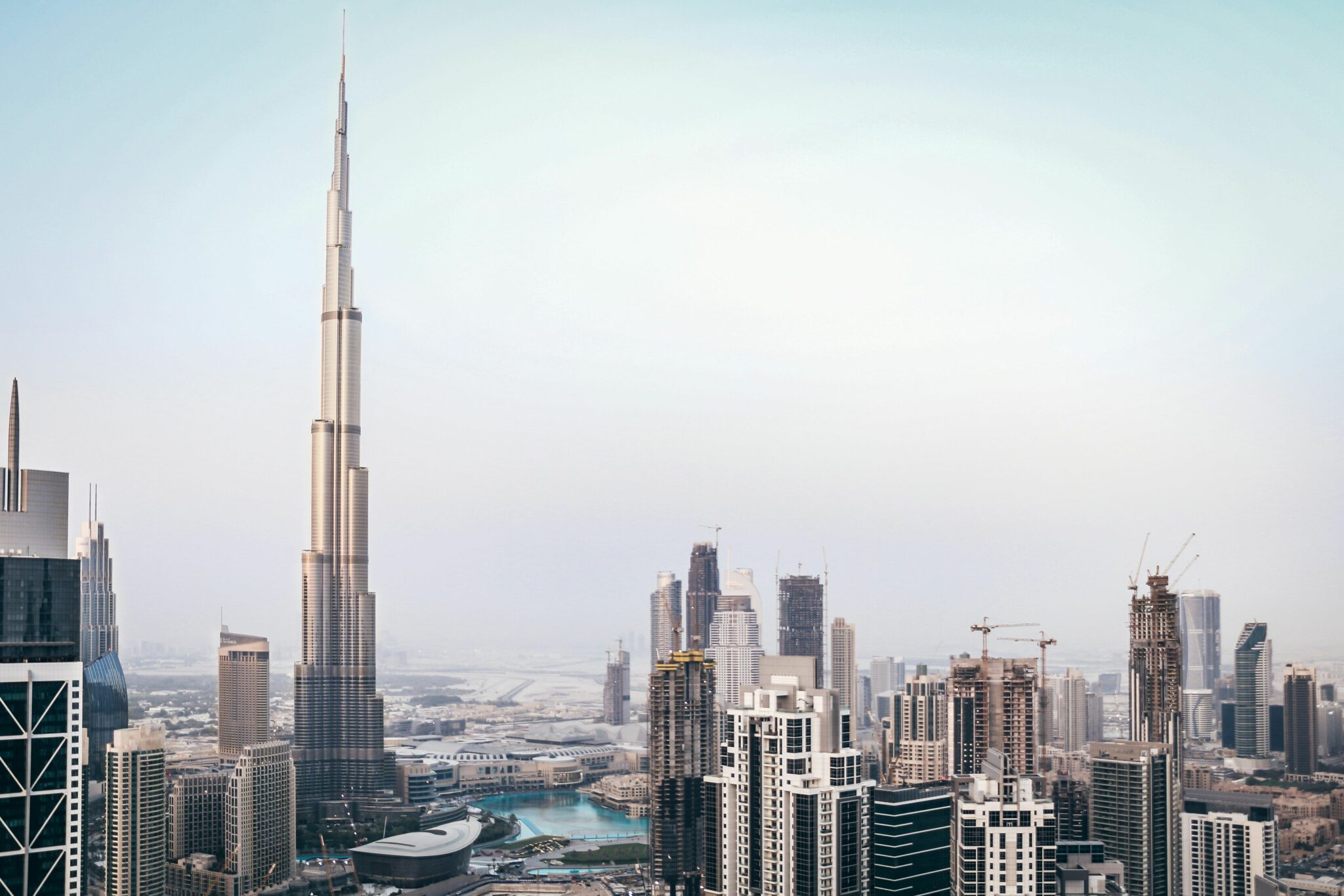 Skyline of Dubai featuring the towering Burj Khalifa surrounded by modern high-rises and construction cranes under a clear, pale blue sky.