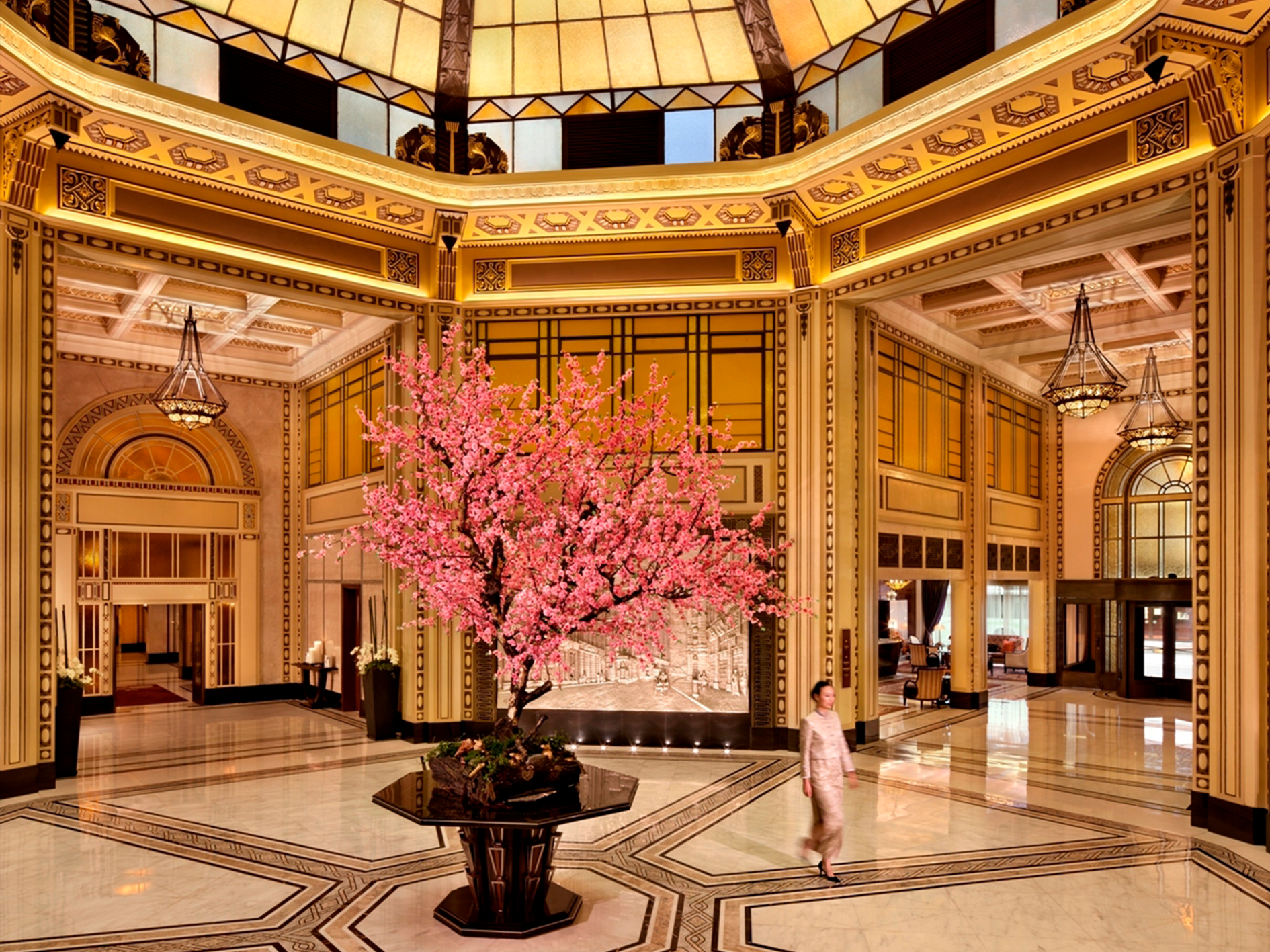 Luxurious hotel lobby adorned with a large, blooming pink cherry blossom tree. Elegant woman in traditional attire walks beneath a grand stained-glass dome.