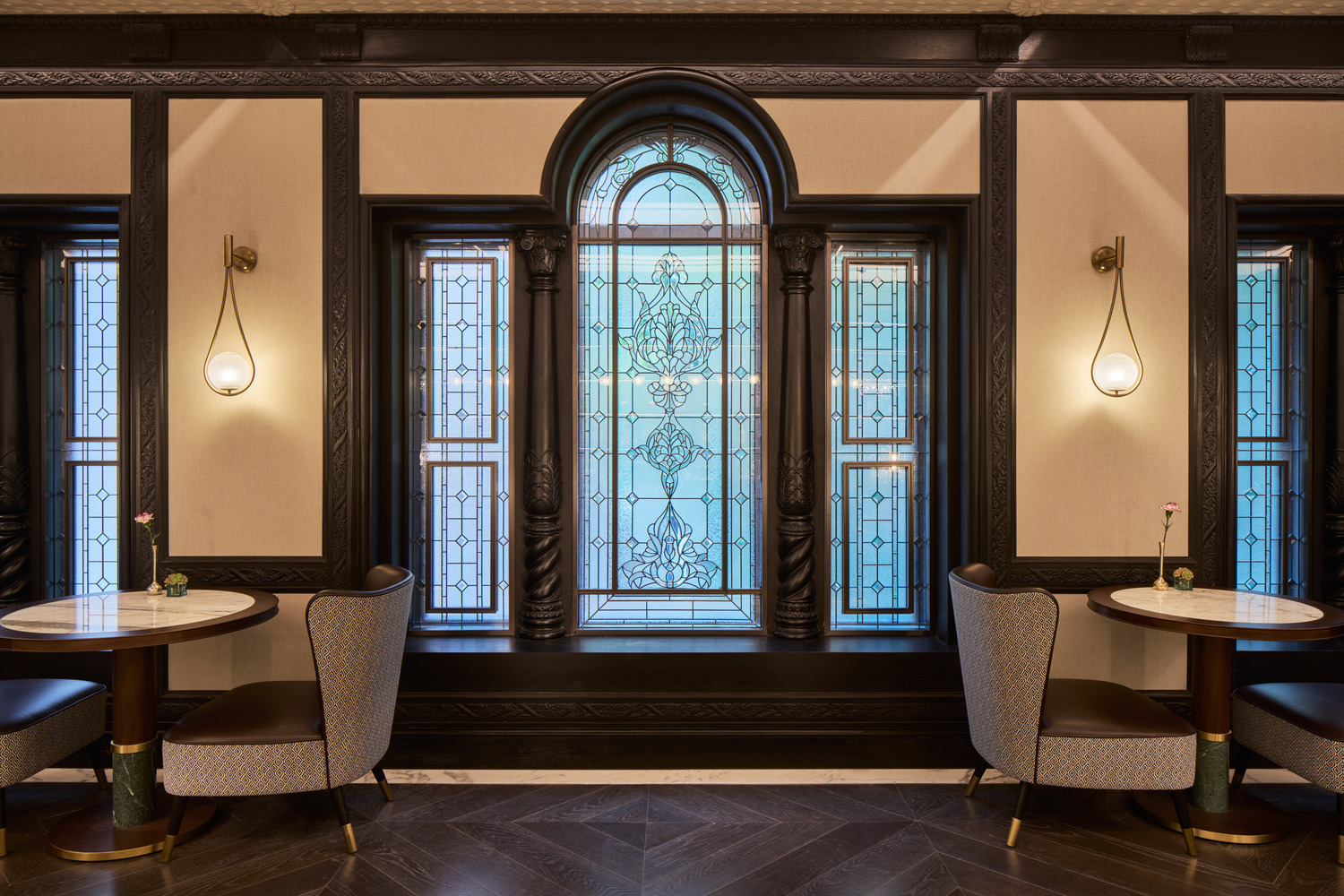 Cafe-style seating area with marble-top tables, upholstered chairs, and tall stained glass windows framed by dark carved wood.