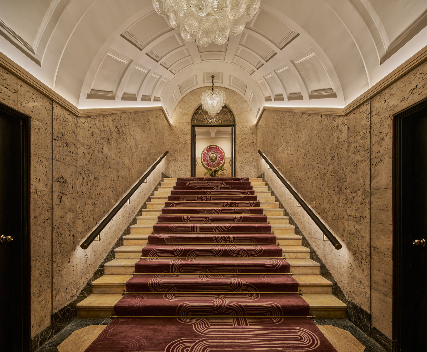 Dramatic staircase with vaulted ceiling, marble walls, crystal chandeliers, and patterned burgundy carpet.