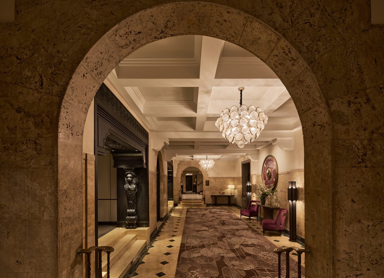 Elegant lobby corridor framed by stone arches, patterned carpet, crystal chandeliers, and classical detailing.