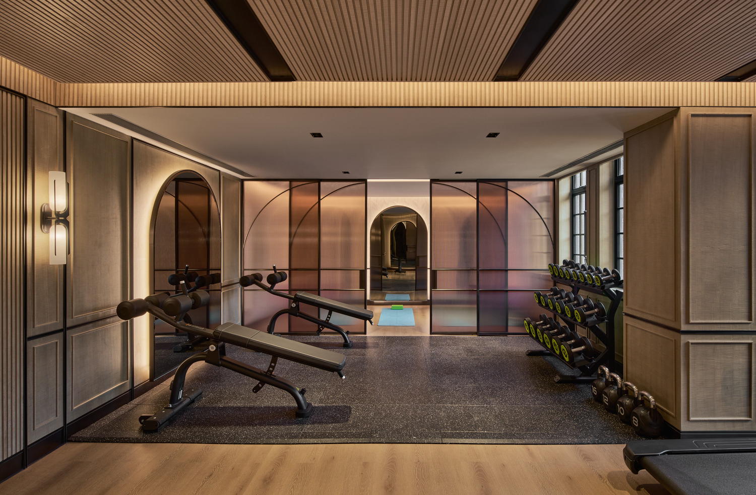 Luxury fitness studio with arched glass partitions, warm wood finishes, free weights, and modern exercise benches.