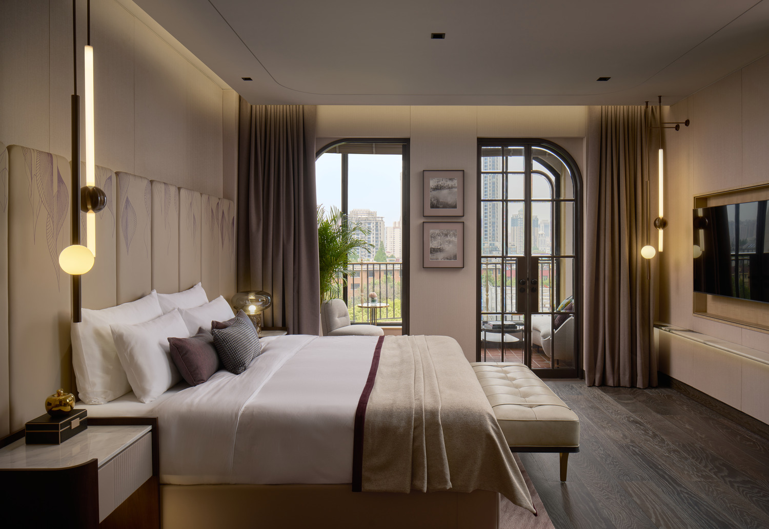 Elegant bedroom with upholstered headboard, pendant bedside lights, floor-to-ceiling curtains, and balcony view of city skyline.
