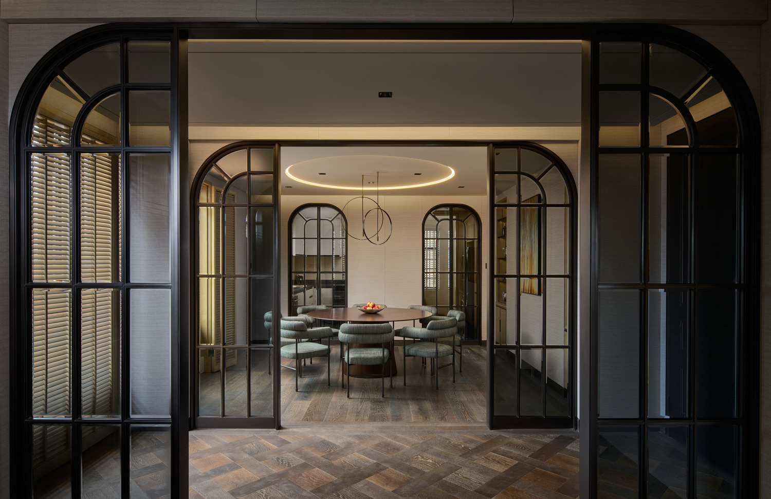 Contemporary dining room framed by glass-panel arched doors, featuring a round wooden dining table, green upholstered chairs, and modern ring chandelier.