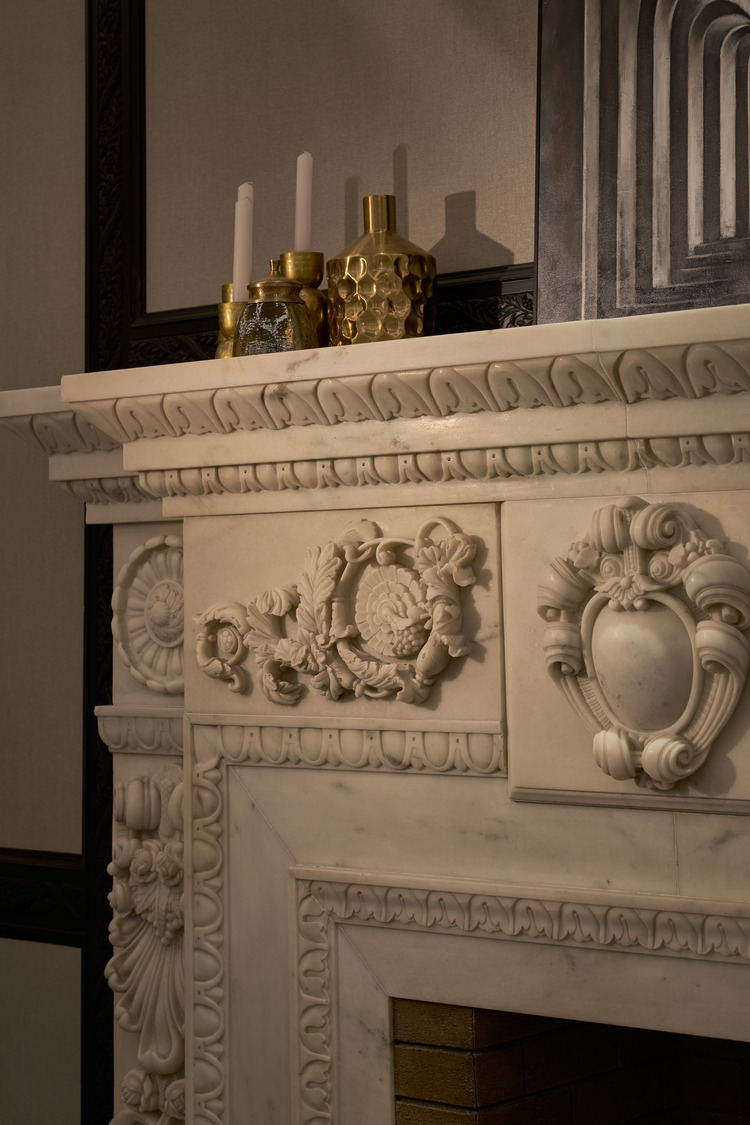 Detail of ornate carved marble fireplace with classical motifs, adorned with gold vases and candles.