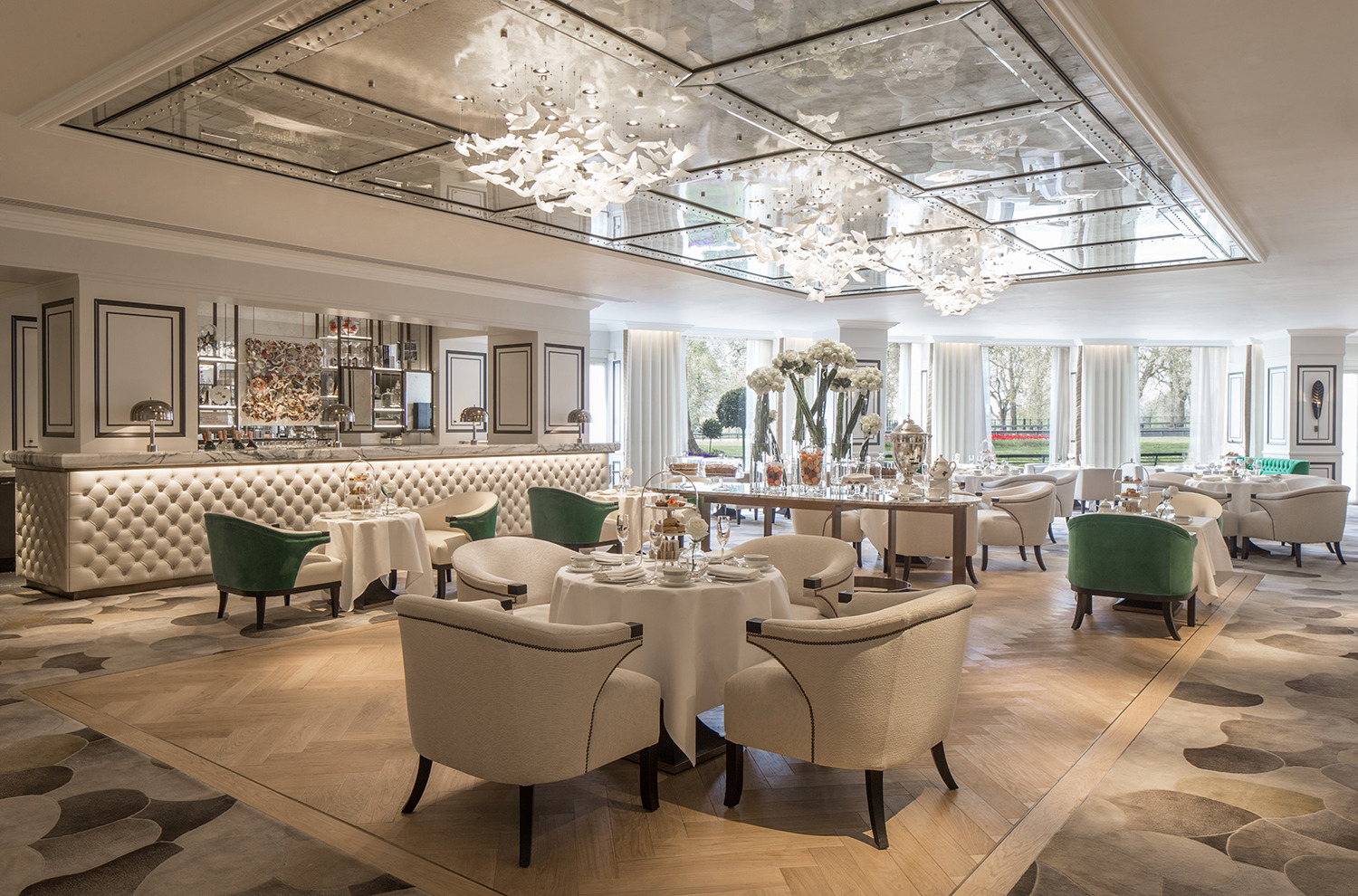 Elegant restaurant interior with tufted banquette seating, round tables set for afternoon tea, and striking mirrored ceiling with sculptural chandeliers.