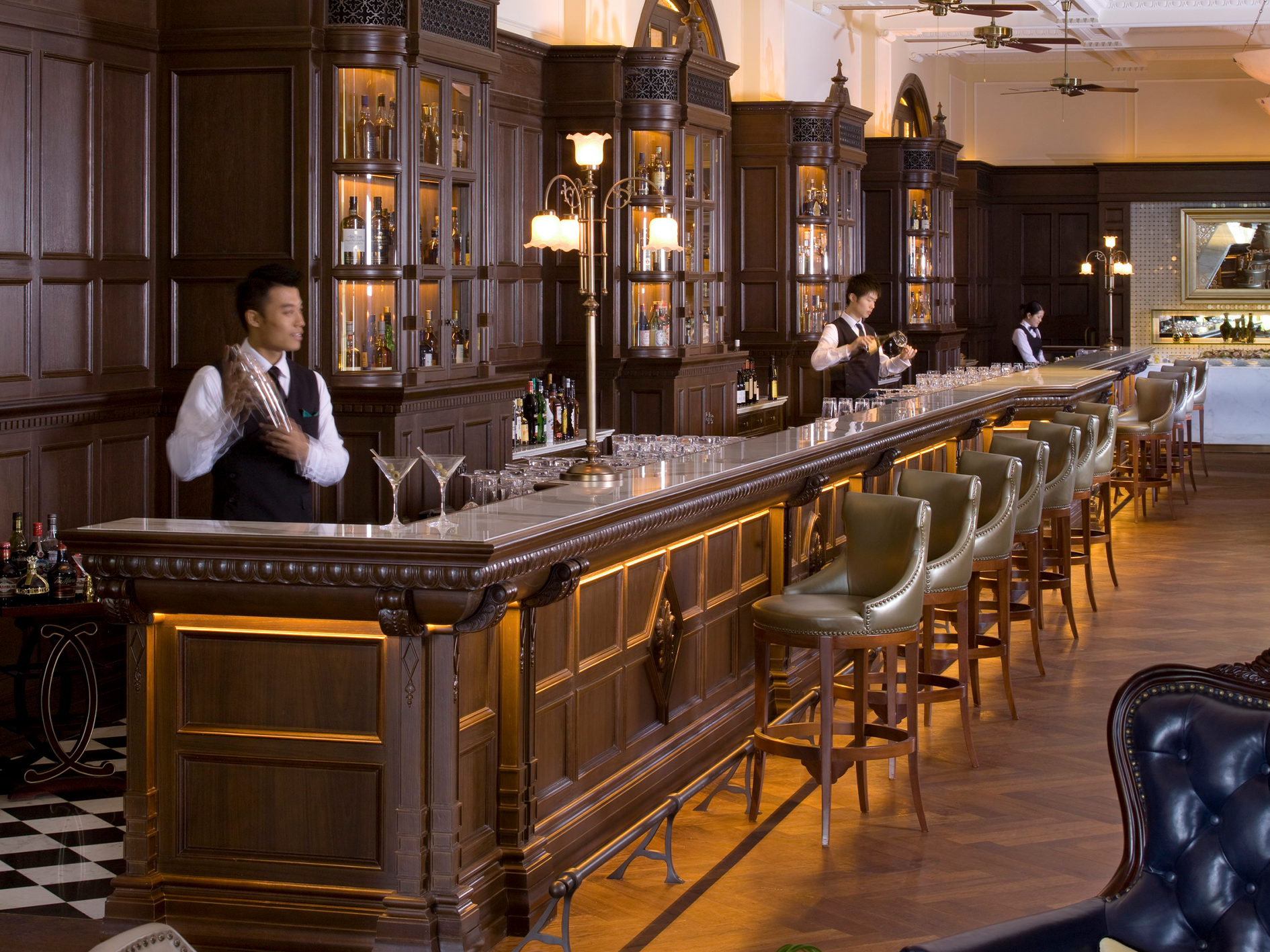 Elegant hotel bar with dark wood paneling, ornate cabinets, and classic bar stools. Bartenders in formal attire prepare drinks under soft, warm lighting.