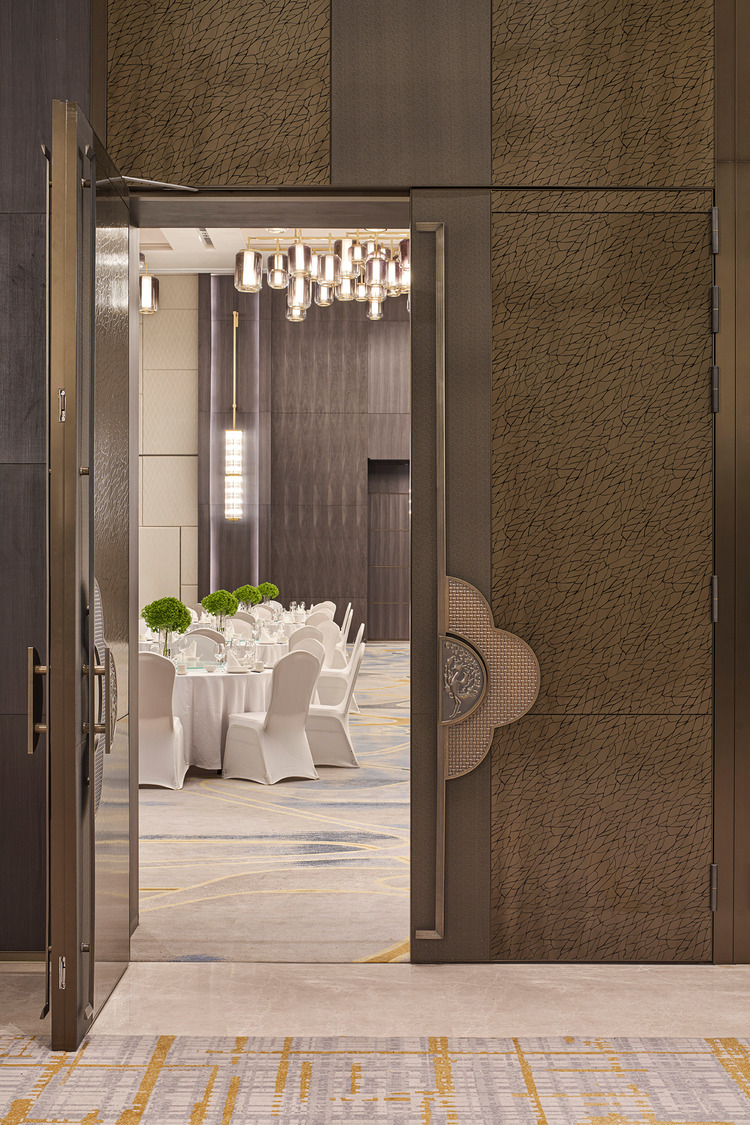 Elegant hotel lobby with modern decor, featuring marble floors, plush seating, and intricate ceiling lights. Soft lighting and greenery add warmth.