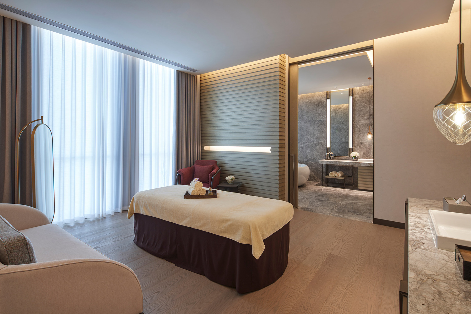 Cozy spa treatment room with two massage beds adorned with white towels and plants. A large window lets in natural light, illuminating the soft, neutral tones.