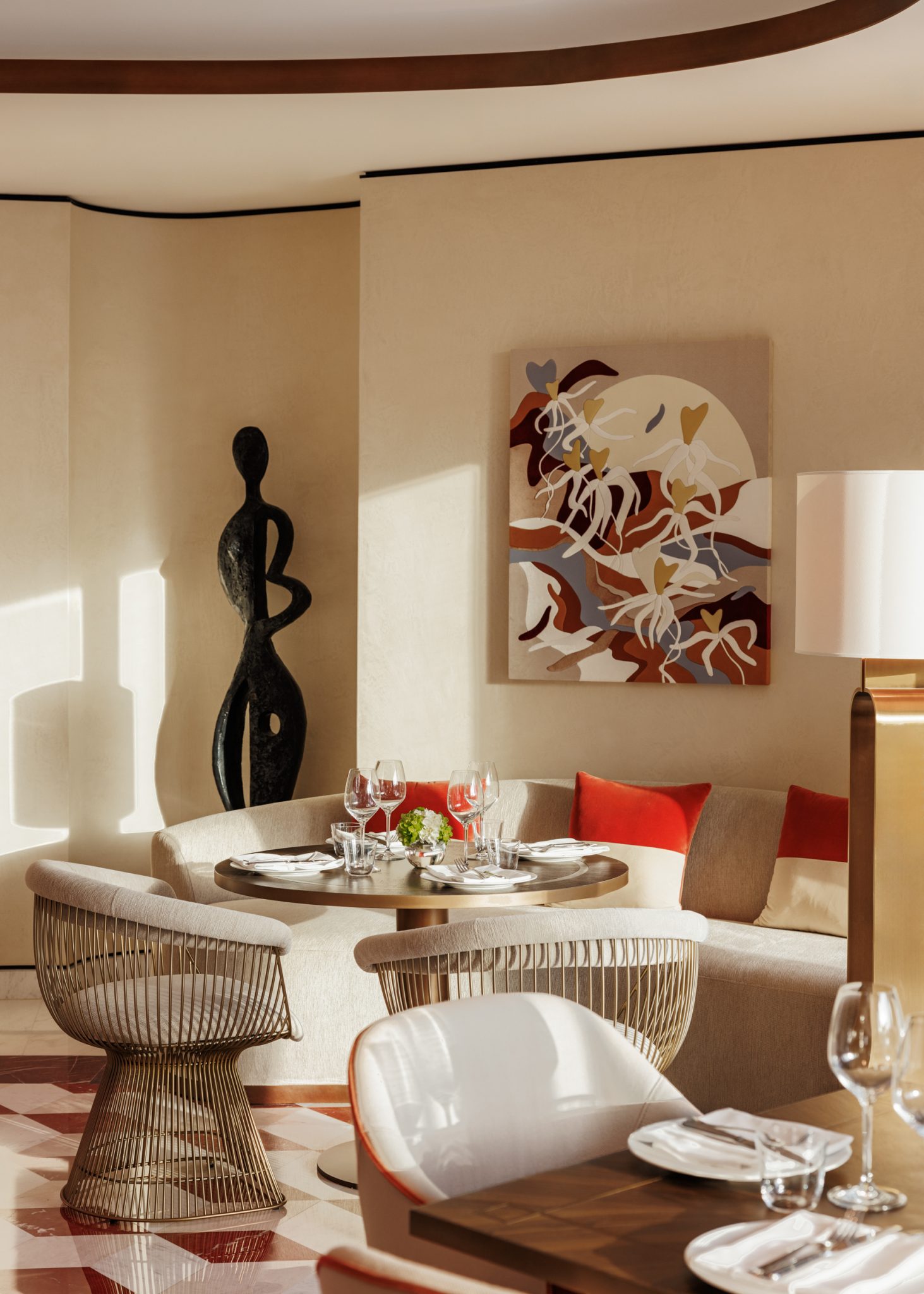 Cozy seating area of a hotel lounge with two beige chairs, a round table, and a red ottoman. A red bookcase displays art and decor. Sunlight streams in from large windows.