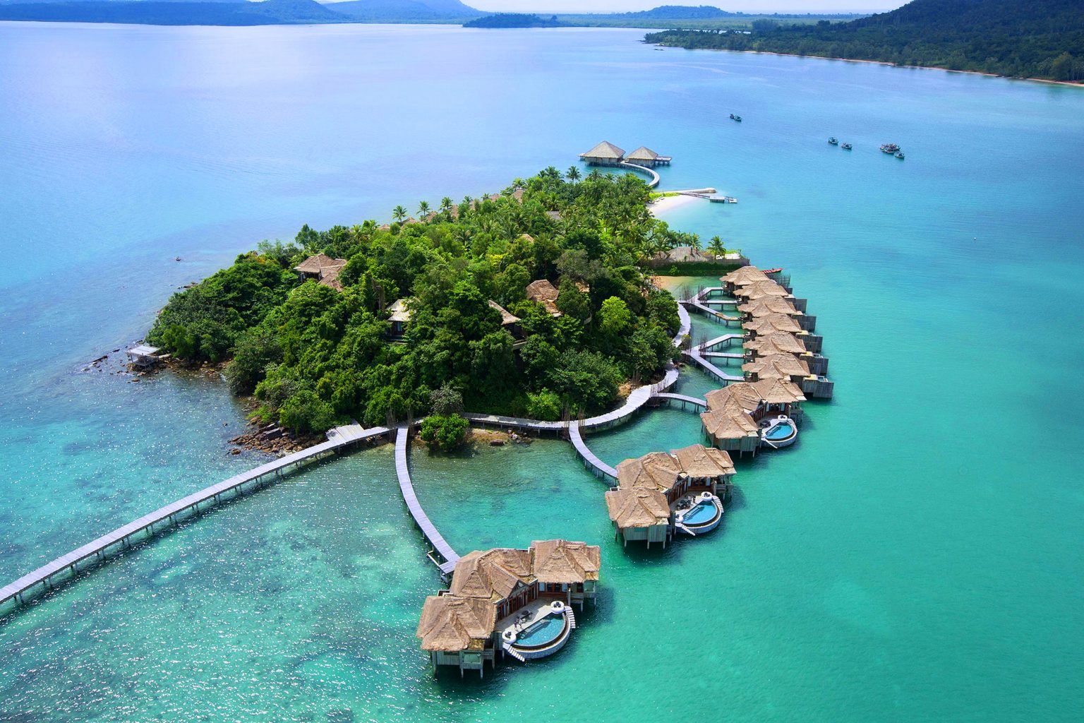 Aerial view of a serene beach setting with palm trees and a large, curved wicker structure on white sand. Lounge chairs and a table are arranged nearby.