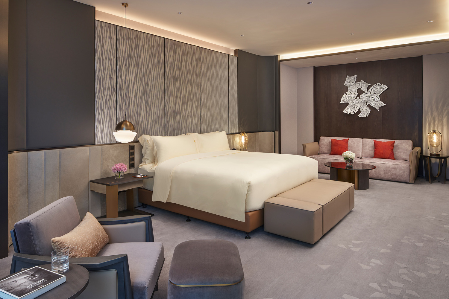 Modern hotel room with a plush white bed, dark wood accents, large TV, artistic wallpaper, and an elegant shelf. Warm lighting creates a cozy ambiance.