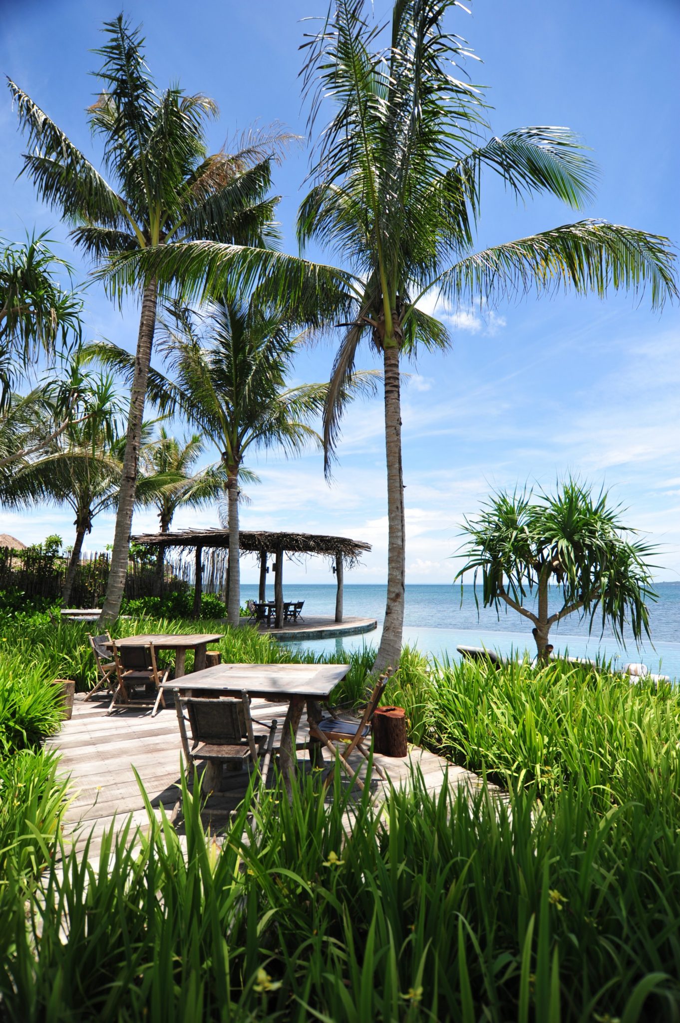 View of a serene beach setting with palm trees and a large, curved wicker structure on white sand. Lounge chairs and a table are arranged nearby.