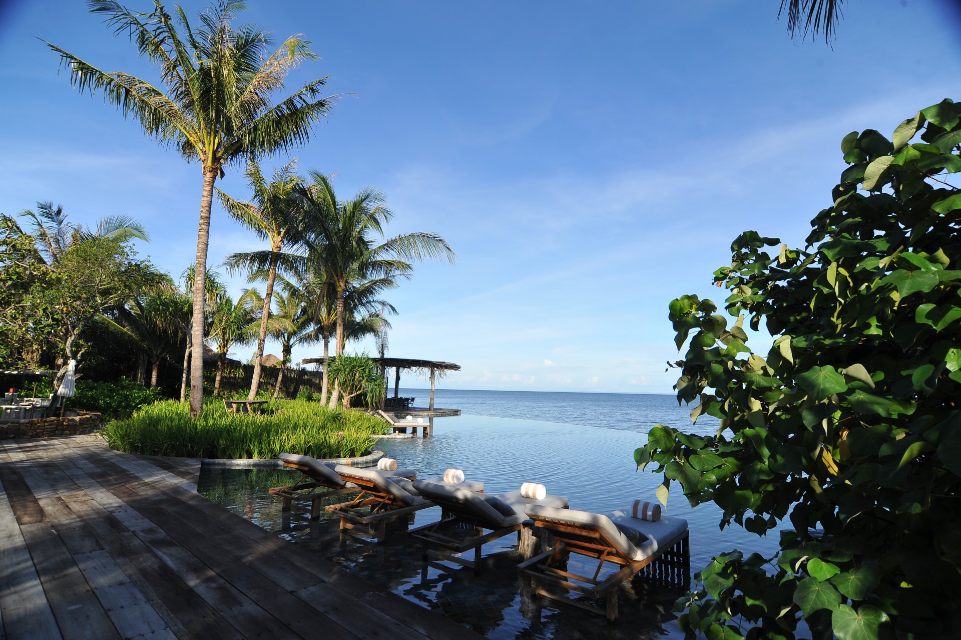View of a serene beach setting with palm trees and a large, curved wicker structure on white sand. Lounge chairs and a table are arranged nearby.