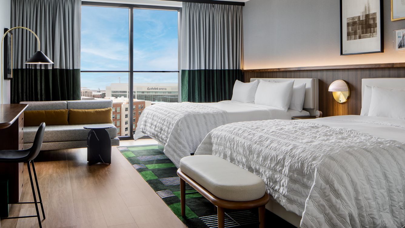 Modern hotel room with two beds, sleek gray tones, and elegant lighting. Features include a seating area, TV, and minibar, creating a cozy yet sophisticated ambiance.