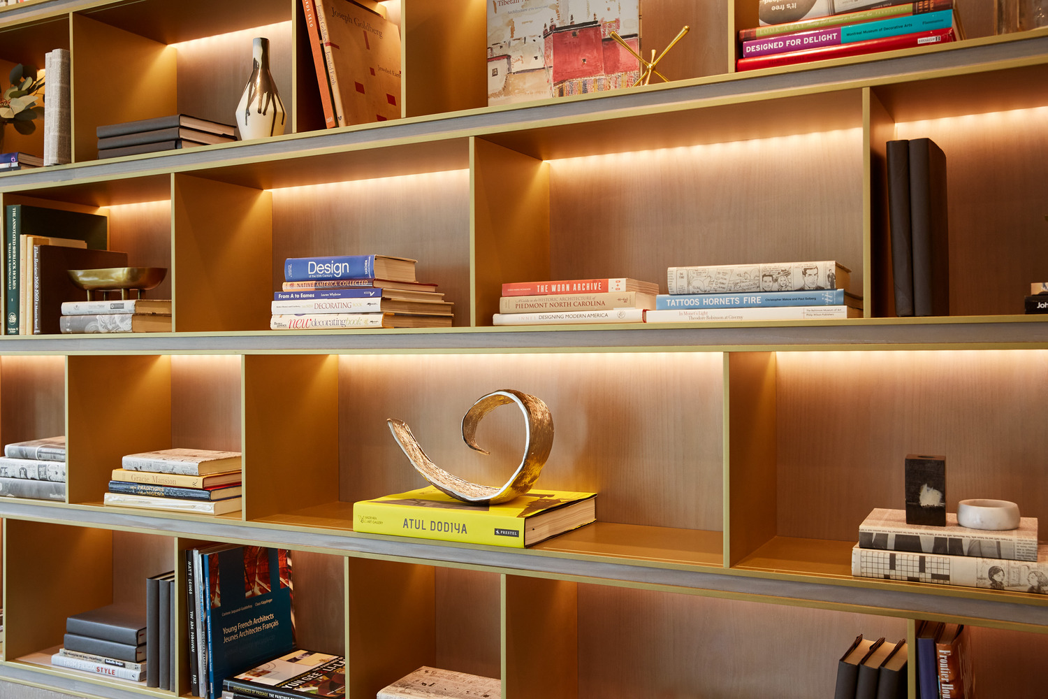 Light-up wooden bookshelf detail with integrated warm lighting illuminating curated books and decorative objects set against natural timber shelving, highlighting grain texture and refined joinery.